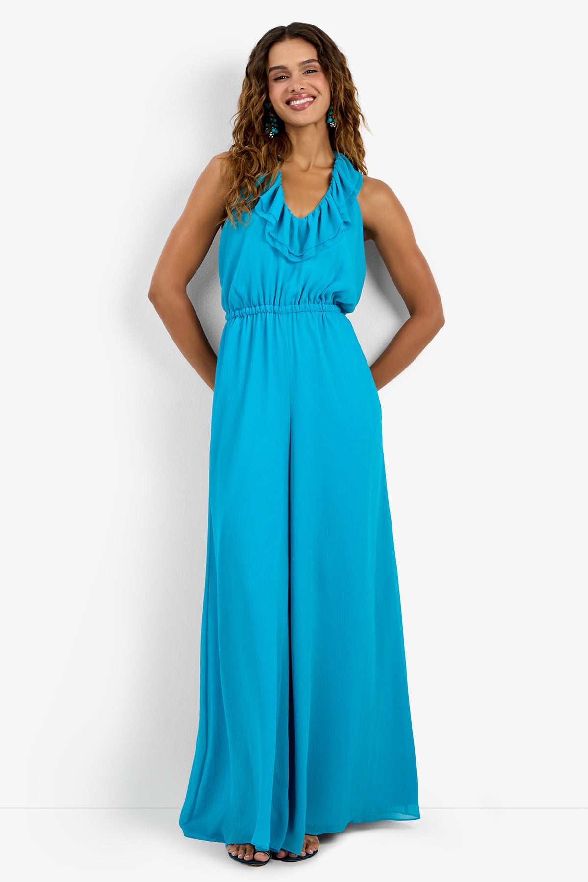 A woman smiles with hands in pockets, wearing the JACKSONVILLE JUMPSUIT—a sleeveless, floor-length, bright turquoise halter-neck jumpsuit in 100% polyester with a ruffled neckline and wide legs. She poses against a white background.