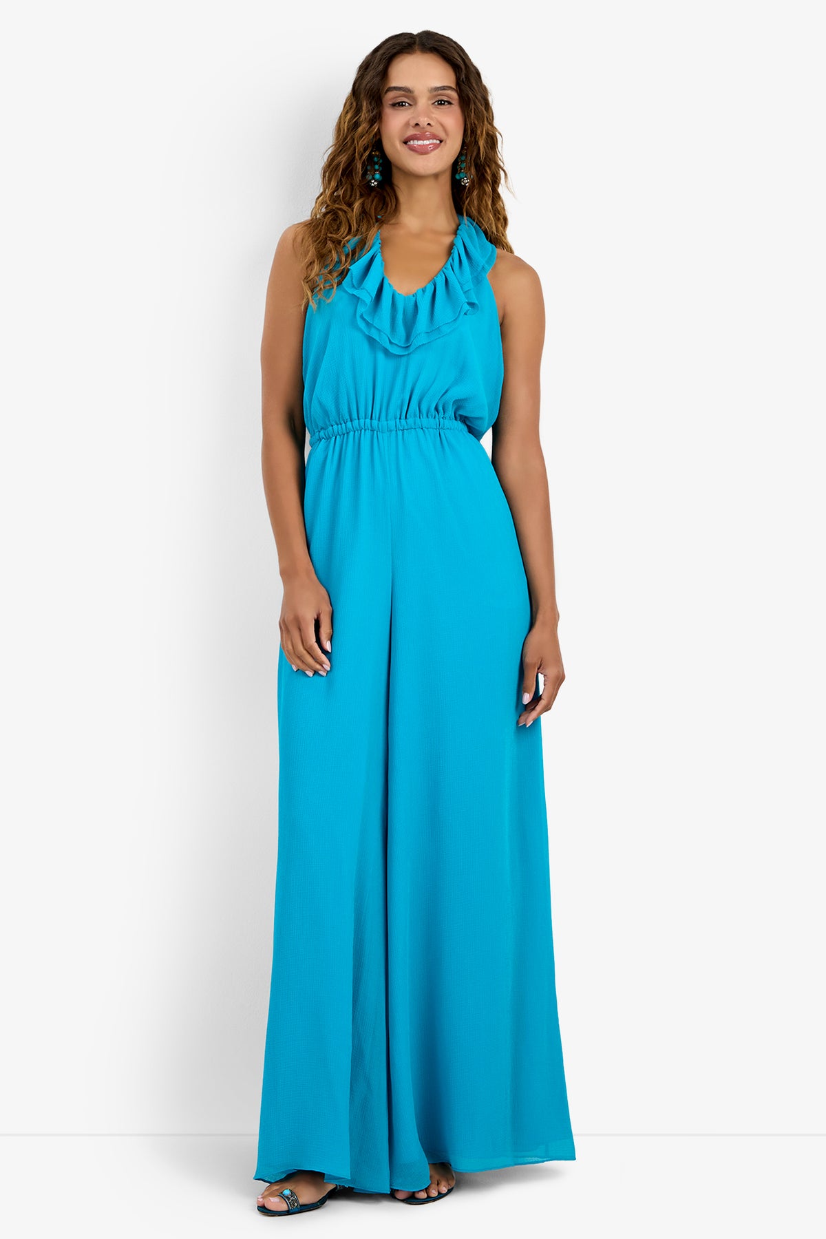 A woman smiles, wearing the JACKSONVILLE JUMPSUIT—a sleeveless turquoise polyester jumpsuit with a ruffled halter neckline and wide-leg pants. She has long wavy hair and open-toe sandals, posed against a plain white background.