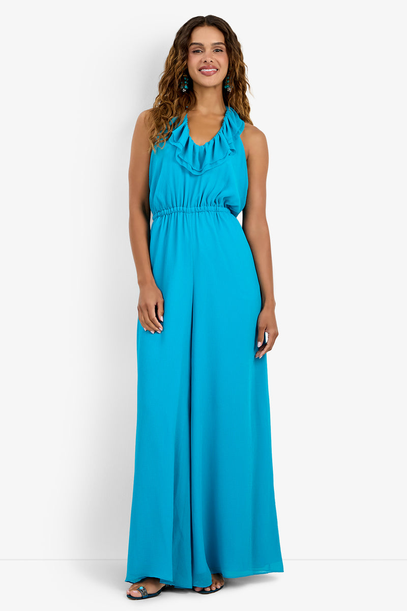 A woman smiles, wearing the JACKSONVILLE JUMPSUIT—a sleeveless turquoise polyester jumpsuit with a ruffled halter neckline and wide-leg pants. She has long wavy hair and open-toe sandals, posed against a plain white background.