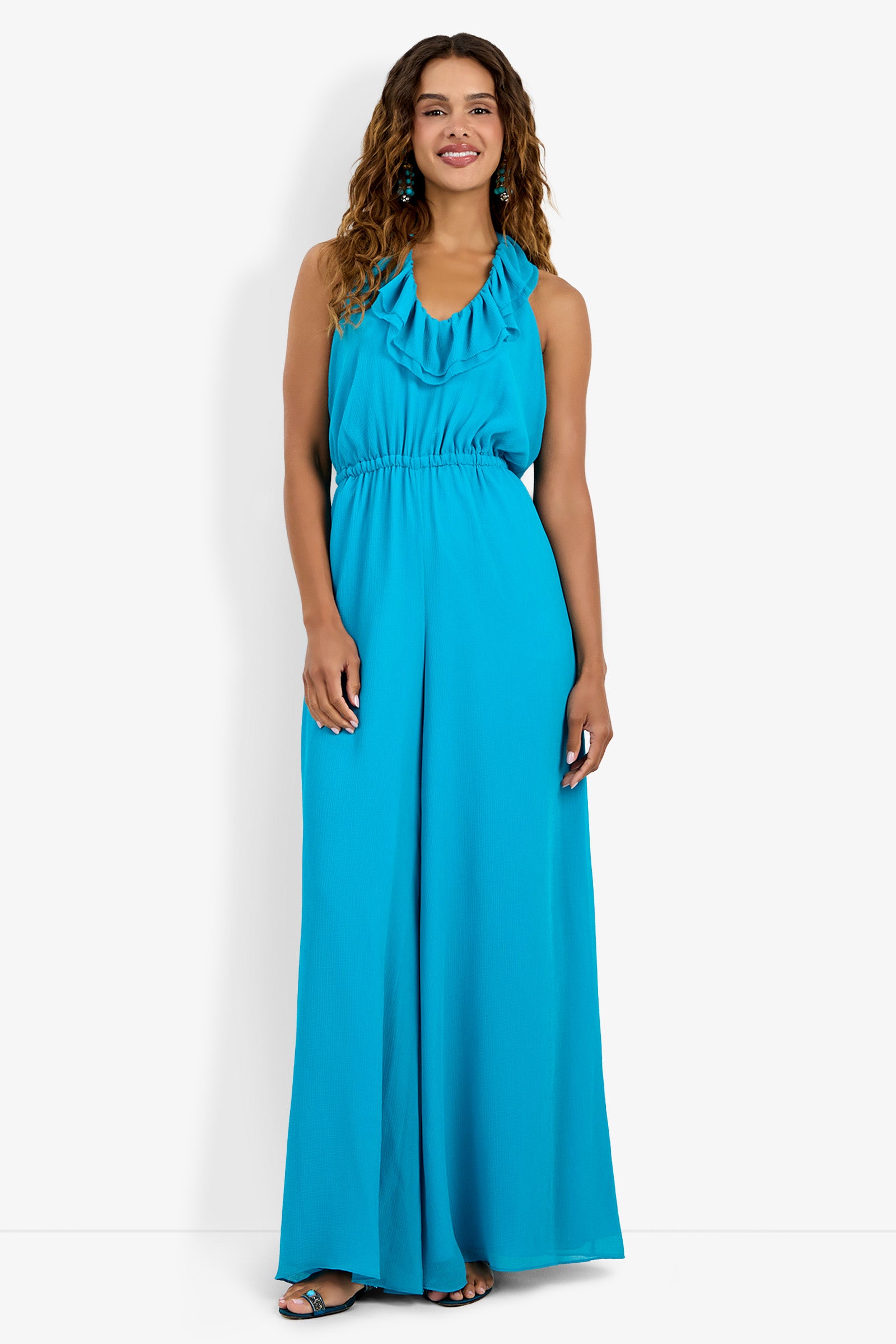 A woman with curly hair smiles in the Jacksonville Jumpsuit—a sleeveless, bright blue halter-neck style with wide-leg pants. She pairs it with black open-toe heels, standing against a plain white background.