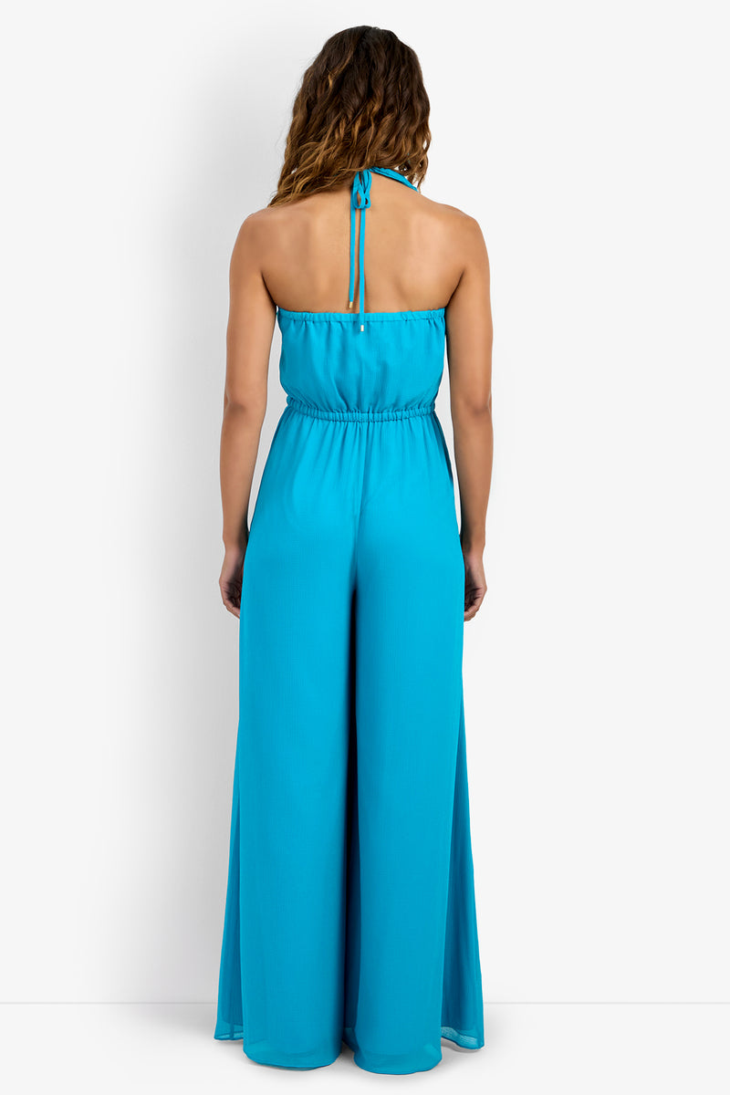A woman with wavy brown hair models the JACKSONVILLE JUMPSUIT—a sleeveless, halter-neck blue jumpsuit made of 100% polyester—standing with her back to the camera against a plain white background.