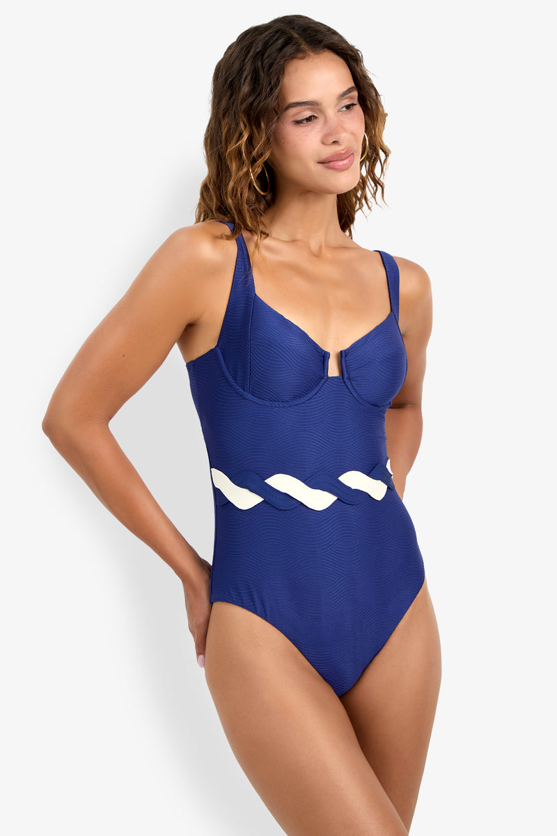 A woman wearing the Underwire One-Piece swimsuit, featuring a blue design with a white wavy accent at the waist, stands against a plain background, looking to the side and smiling gently.