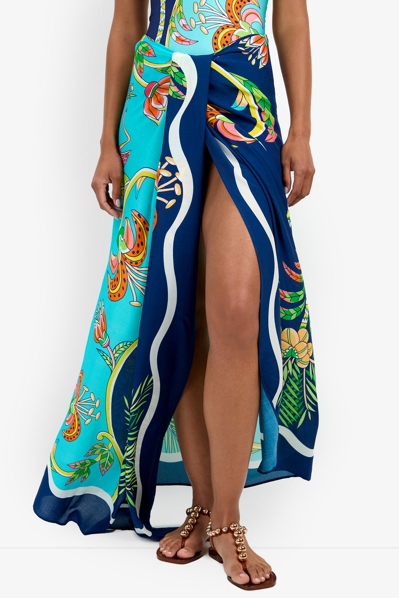 A person wears the Pareo, a brightly colored floral wrap skirt with a high leg slit, paired with gold-accented flat sandals. Only their lower half is visible.