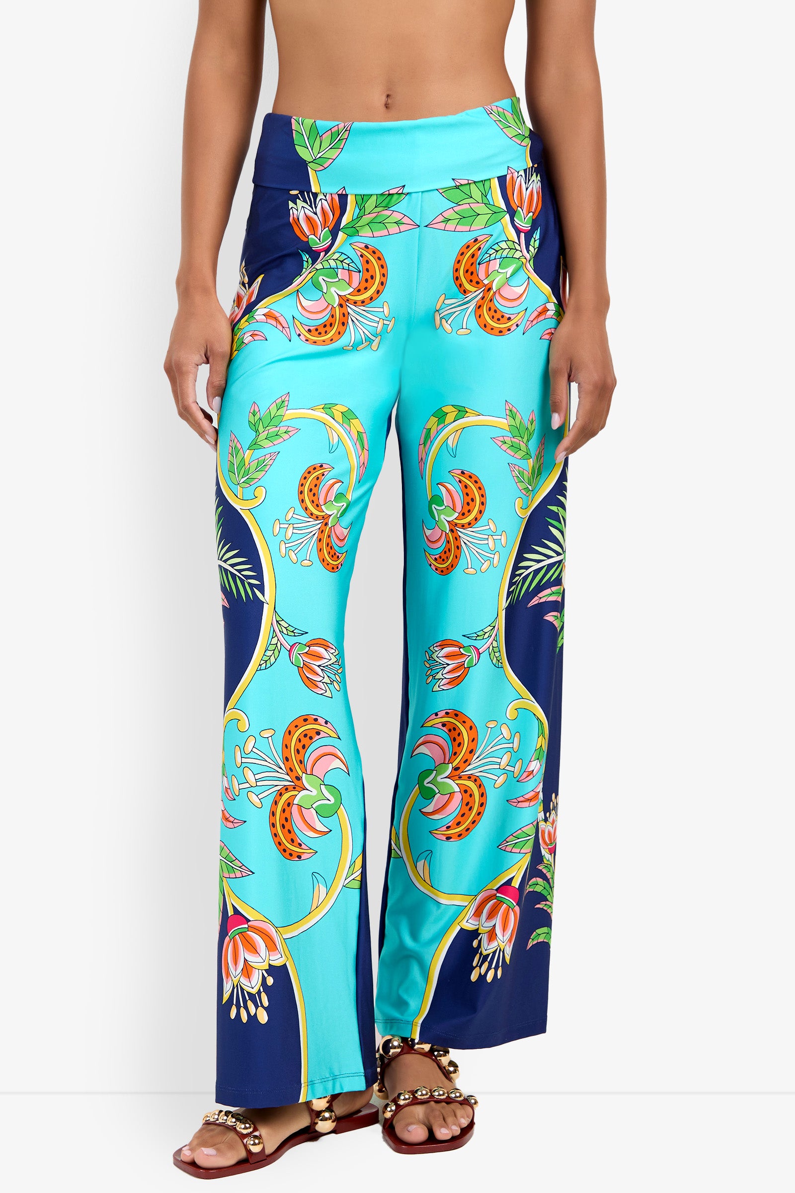A woman models the Swim Pant—high-waisted, wide-leg turquoise pants with a vibrant floral and leaf print—paired with brown beaded sandals, shown from midriff to feet against a white background.