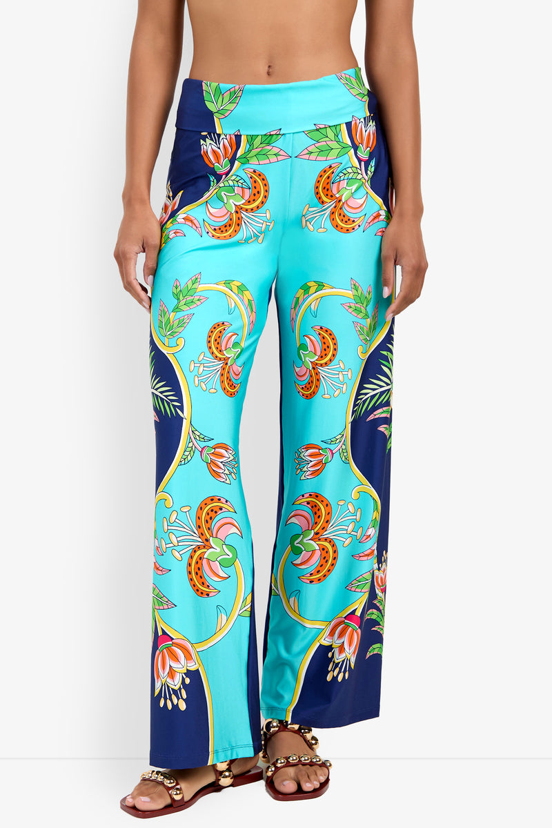 A woman models the Swim Pant—high-waisted, wide-leg turquoise pants with a vibrant floral and leaf print—paired with brown beaded sandals, shown from midriff to feet against a white background.