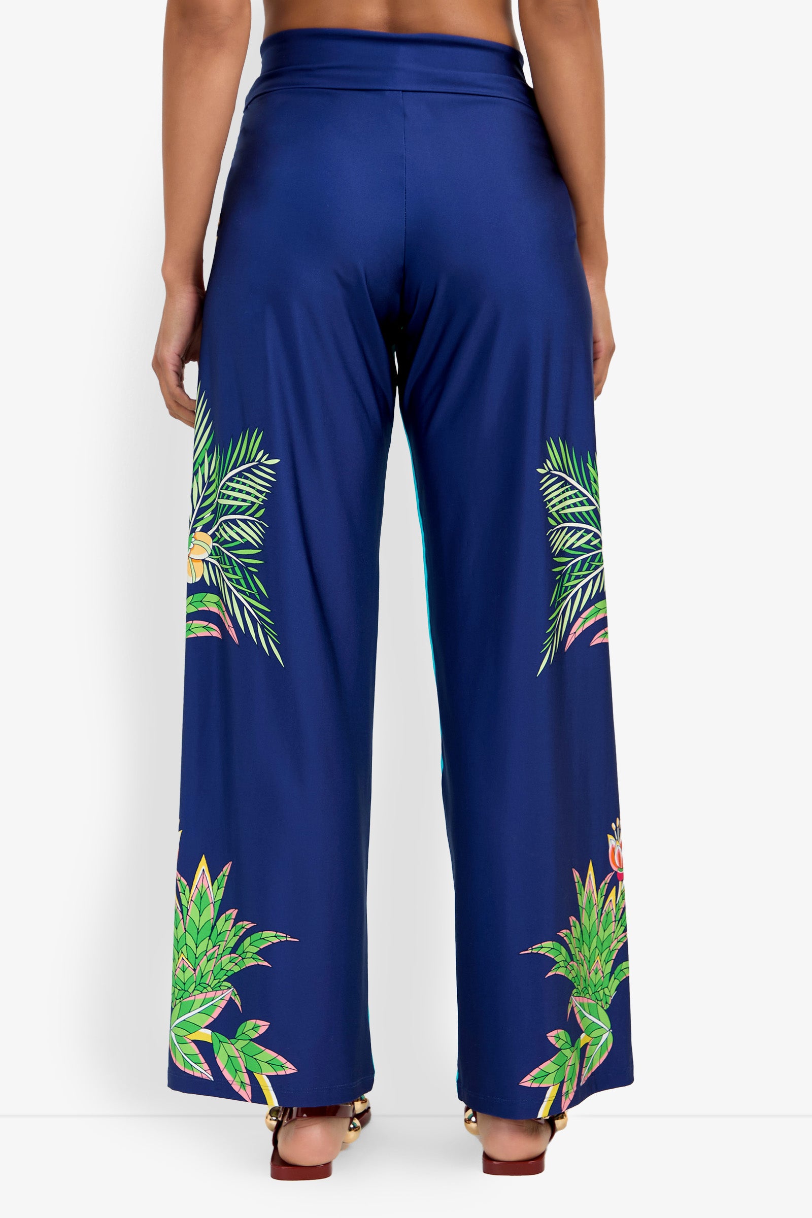 A person wearing Swim Pant—high-waisted, wide-leg blue pants with tropical green leaf and floral prints on the back and sides—stands against a white background.