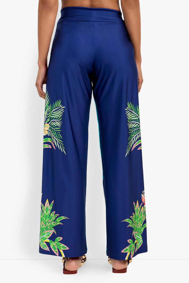 A person wearing Swim Pant—high-waisted, wide-leg blue pants with tropical green leaf and floral prints on the back and sides—stands against a white background.