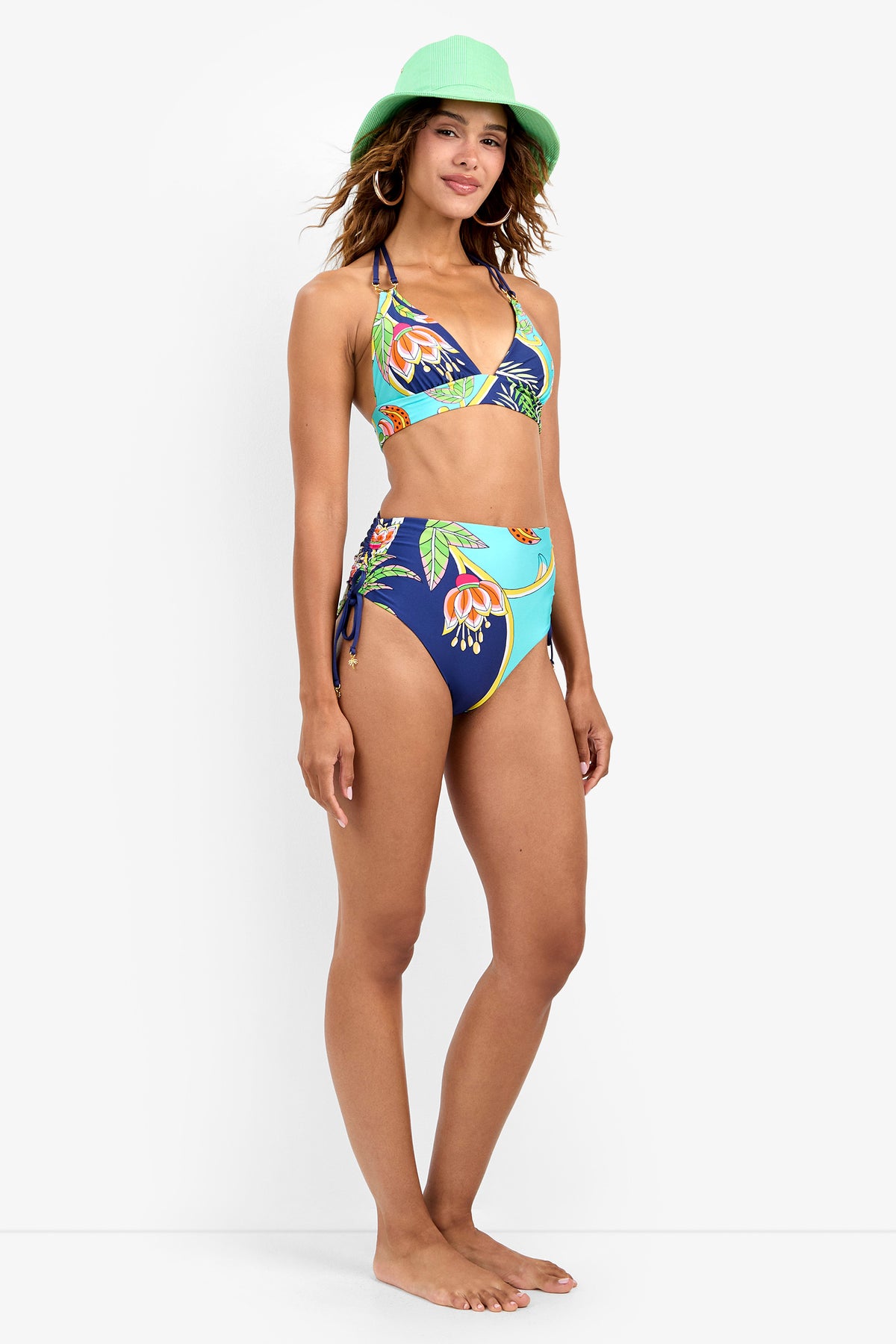 A woman wearing a green hat and a tropical floral bikini with the High Waist Bikini Bottom stands barefoot, smiling, against a plain white background.