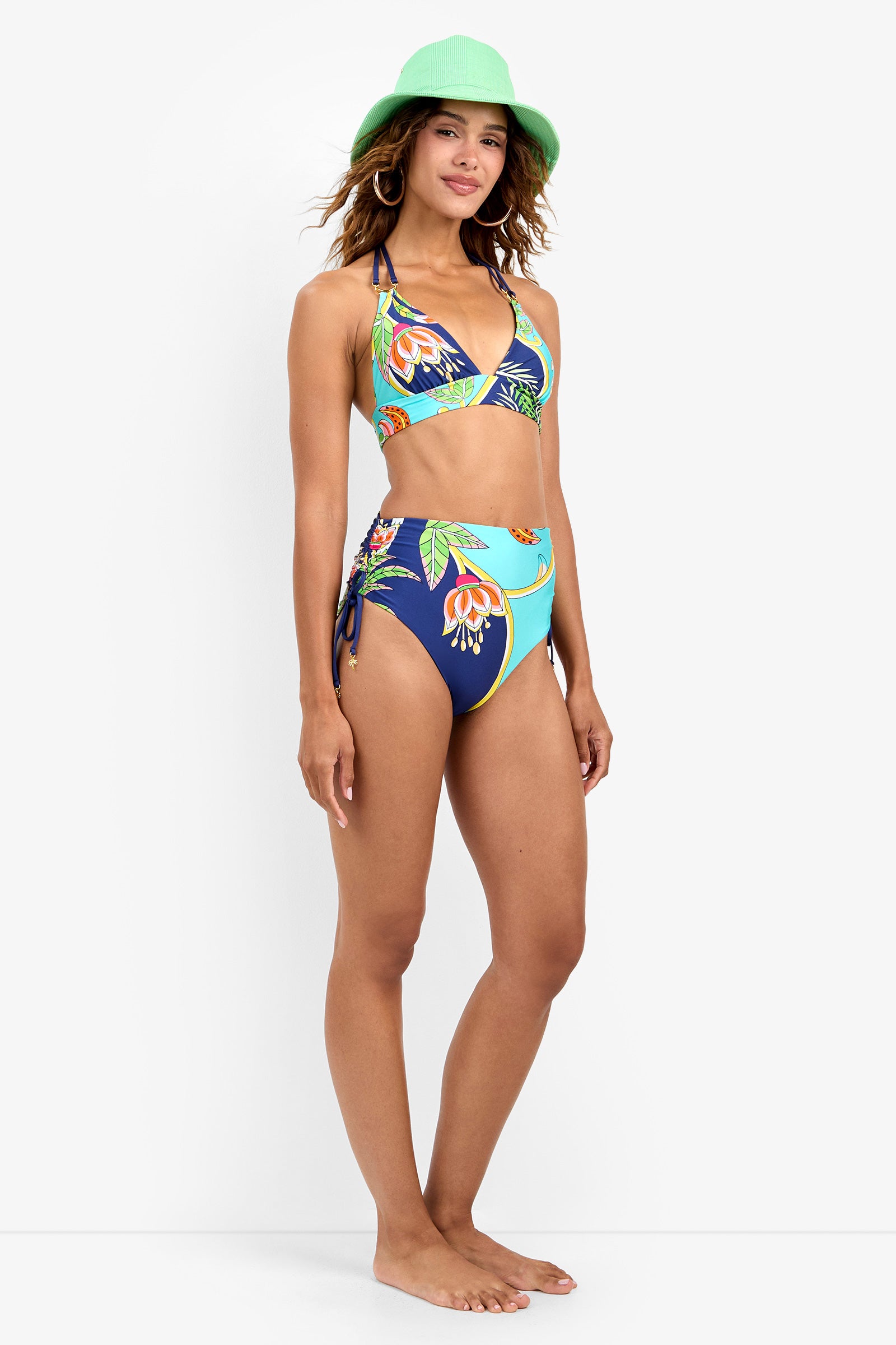 A woman wearing a green hat and a tropical floral bikini with the High Waist Bikini Bottom stands barefoot, smiling, against a plain white background.