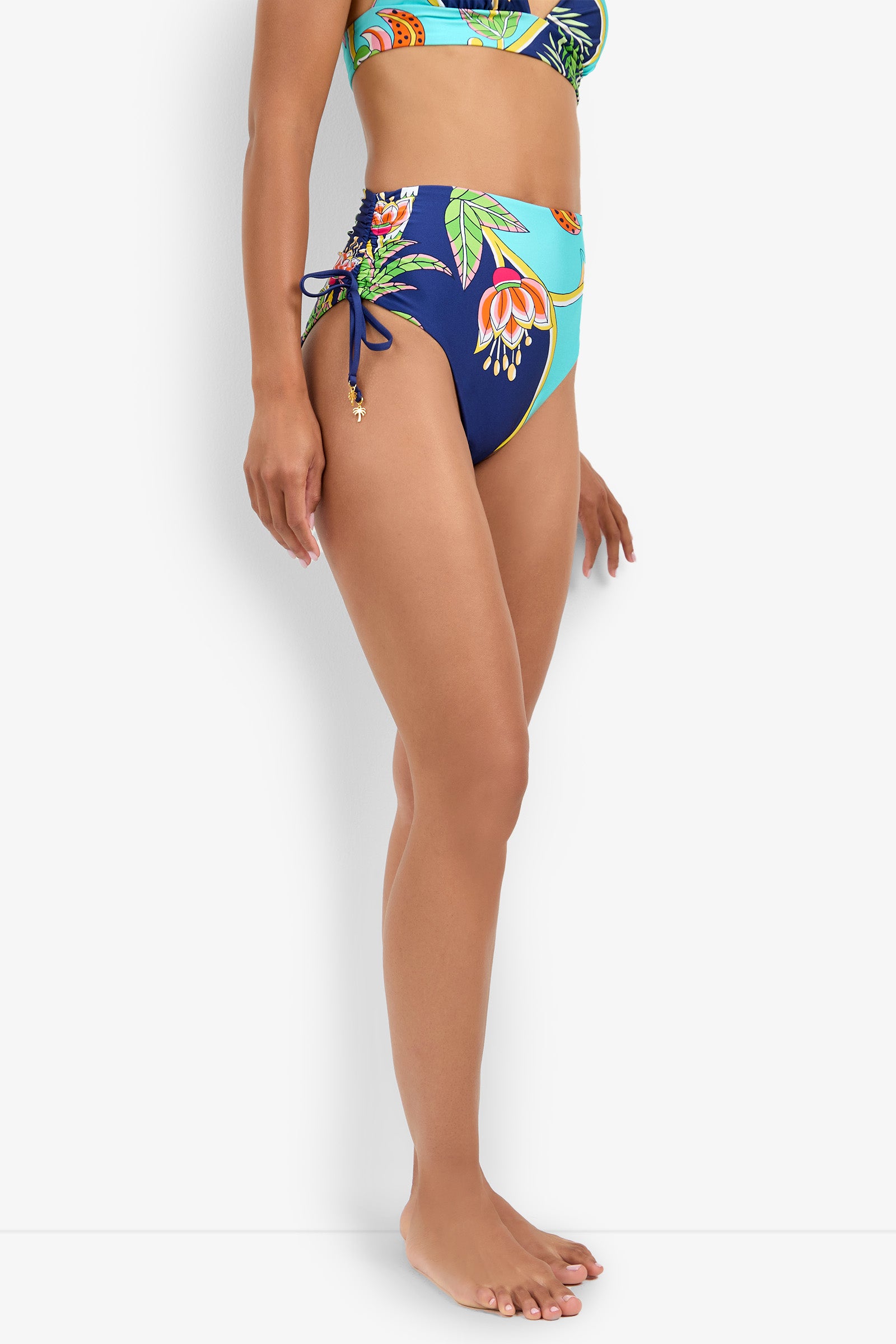 A woman wearing the High Waist Bikini Bottom with a tropical floral print and side-tie details stands barefoot against a white background, facing left, shown from mid-torso to feet.