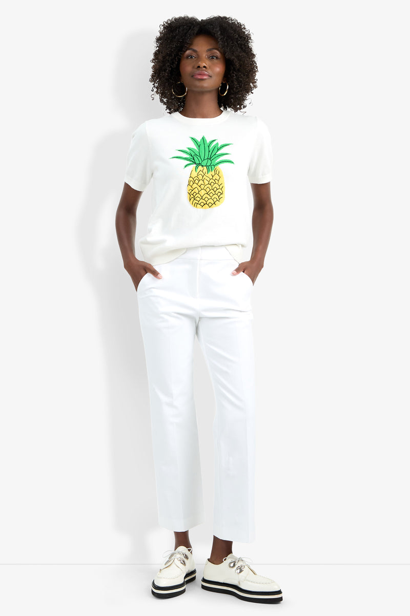 A person with curly hair stands against a white background wearing the Maui Old Fashioned Sweater, white pants, and white platform shoes, with their hands in their pockets.