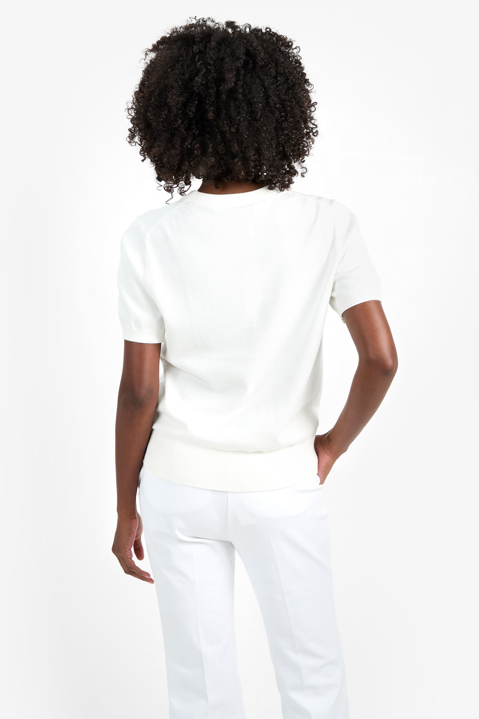 A person with curly hair, wearing the MAUI OLD FASHIONED SWEATER and white pants, stands with their back to the camera against a plain white background.