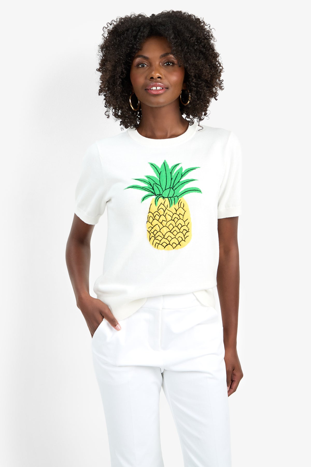 A woman with curly hair wears the MAUI OLD FASHIONED SWEATER, a white short-sleeve top with a large yellow pineapple graphic, paired with white pants. She stands against a plain background, one hand in her pocket and smiling slightly.