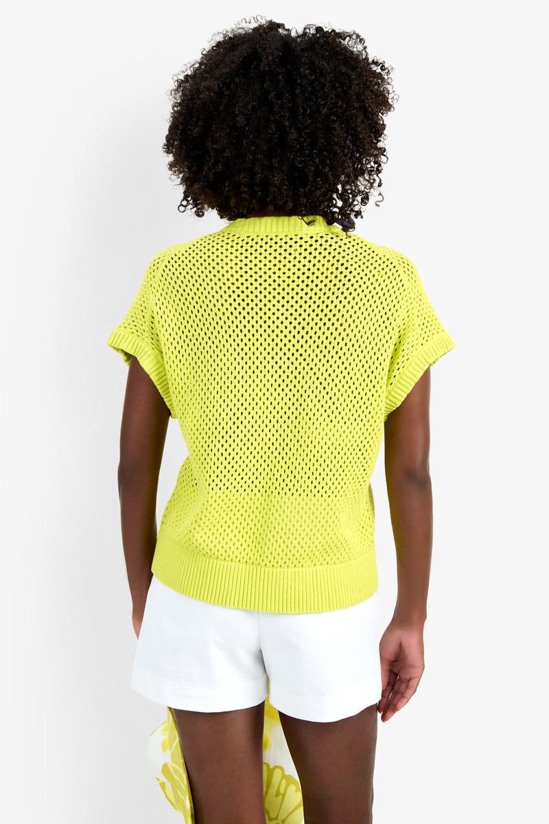 A person with curly hair is seen from the back, wearing the HANA ZIP CARDIGAN—a short-sleeved, yellow mesh knit top made from 100% cotton—and white shorts, set against a plain white background.
