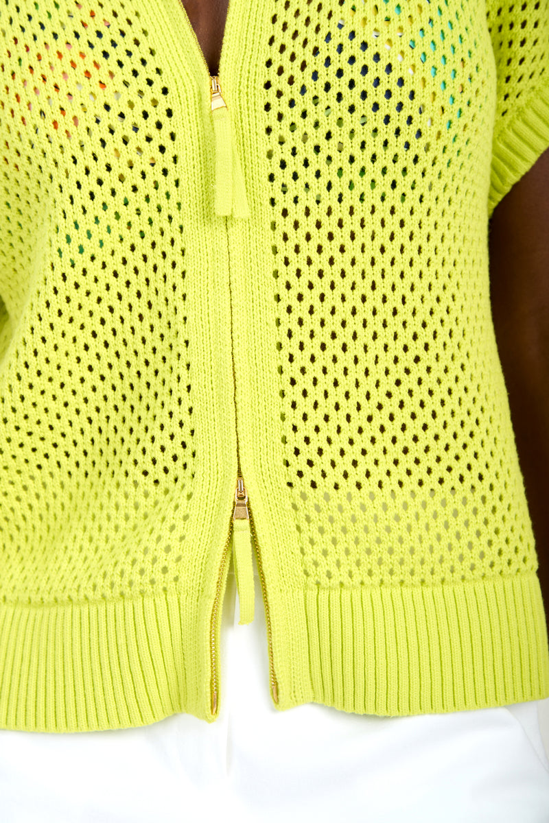 Close-up of a person wearing the HANA ZIP CARDIGAN, a bright yellow-green, short-sleeve, 100% cotton zip-up knit with perforated pattern and ribbed hem, paired with white bottoms.