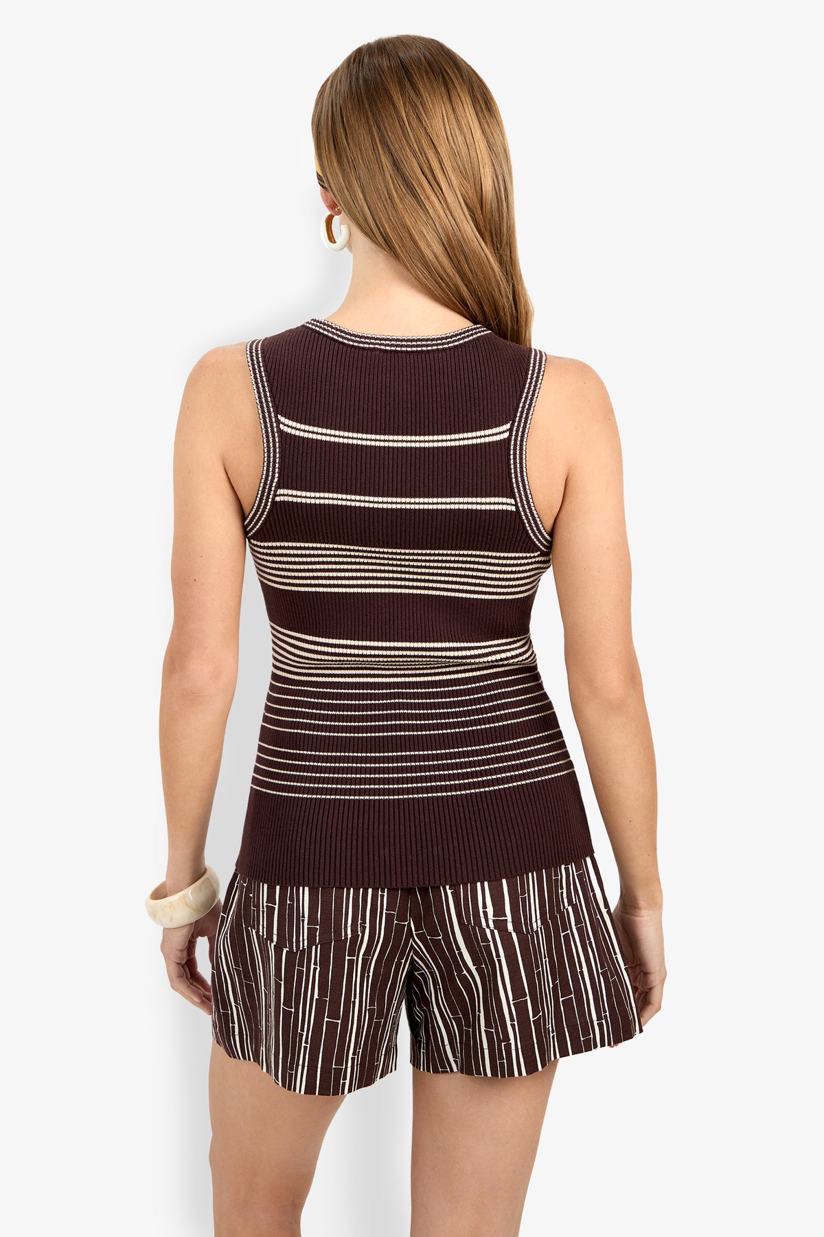 A woman faces away wearing the Paolini Tank—a sleeveless, fitted brown top with white horizontal stripes—paired with matching brown shorts featuring white stripes. She has long hair, hoop earrings, and a chunky bracelet.