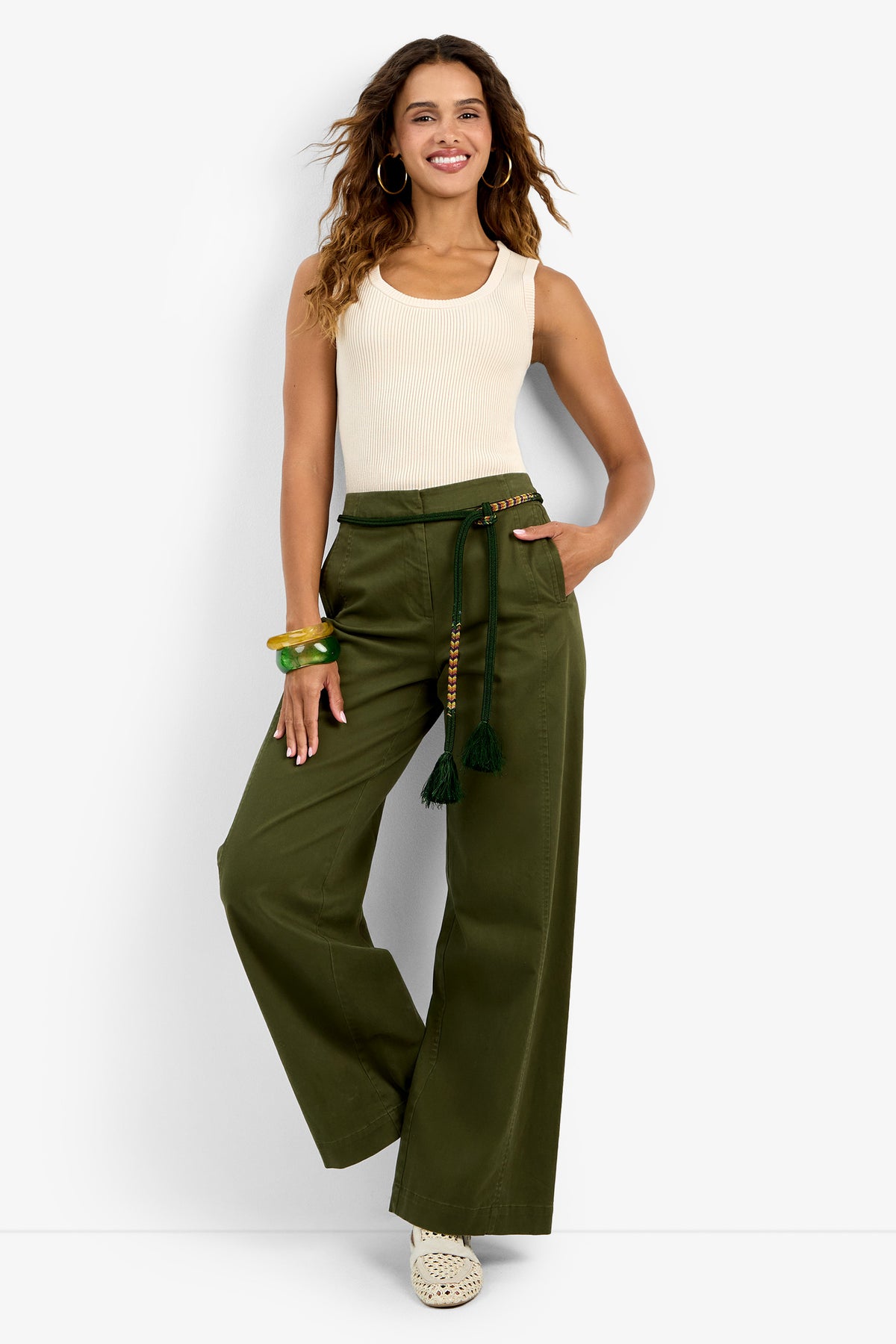 A woman smiles against a white background, wearing the Paolini Tank—a sleeveless cream ribbed top—paired with wide-leg olive green pants with a tasseled belt, green bangles, and white woven shoes.