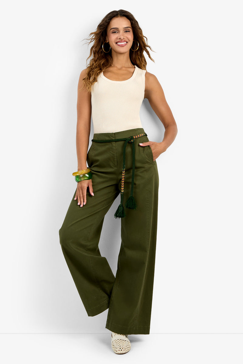 A woman smiles in the Paolini Tank, a sleeveless cream top, paired with high-waisted olive green wide-leg pants featuring a braided belt. She accessorizes with wavy hair, hoop earrings, bangles, and white woven shoes.
