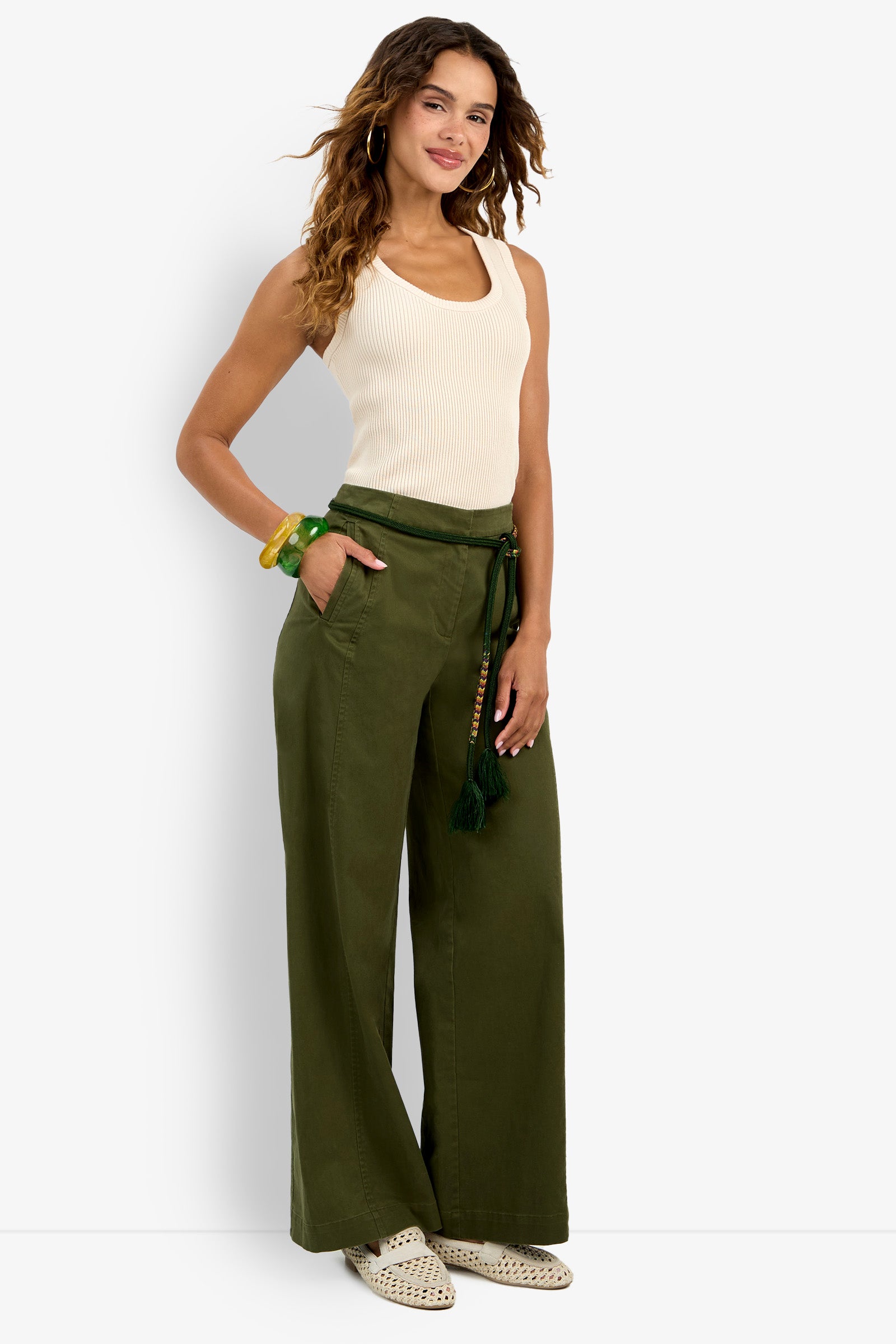 A woman smiles in the white Paolini Tank, olive wide-leg pants with a rope belt, beige woven slip-ons, gold hoop earrings, and green and yellow bangles.