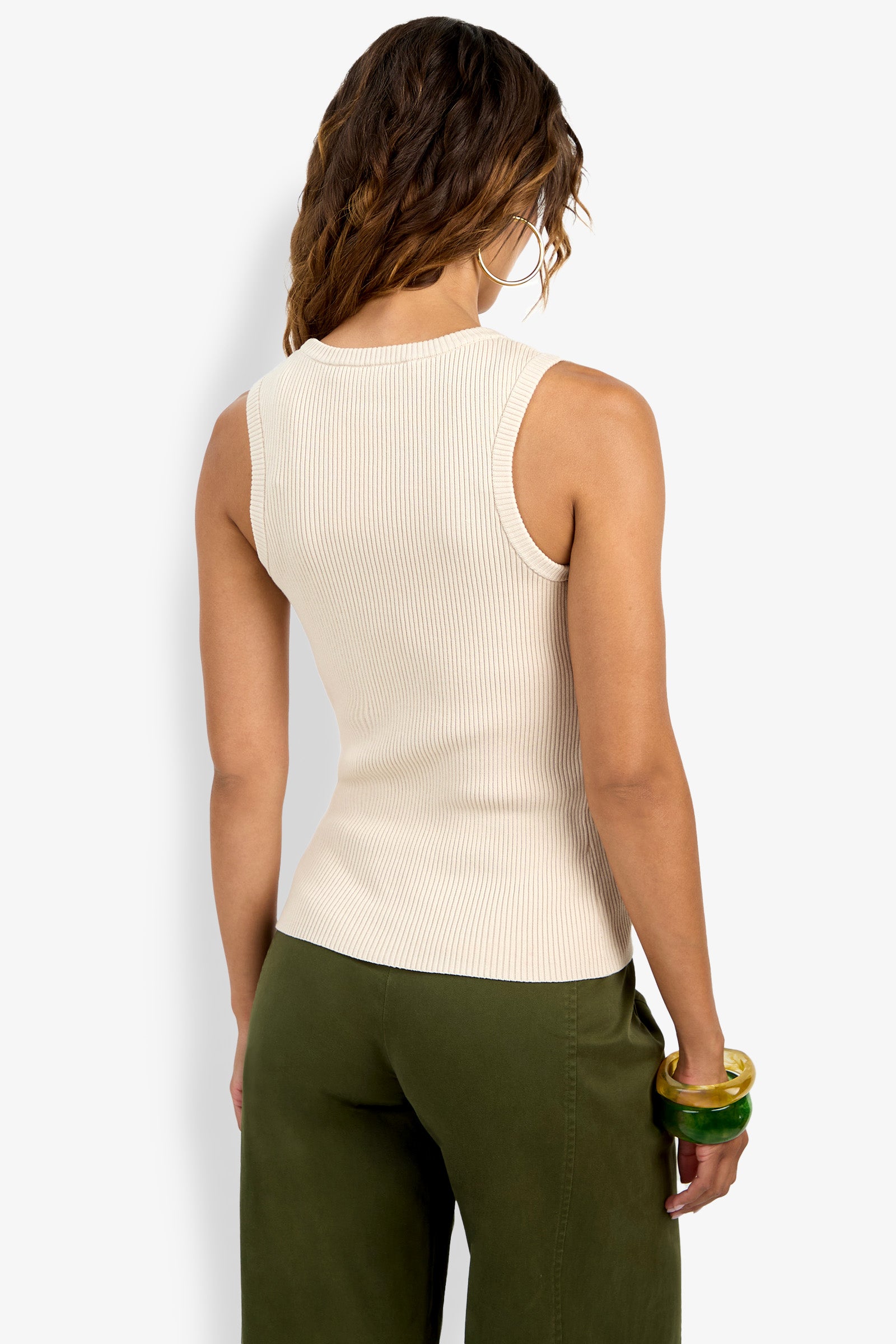 A woman with wavy brown hair is seen from behind, wearing the sleeveless, ribbed Paolini Tank in cream and olive green pants. She accessorizes with large hoop earrings and multiple chunky bangles on her right wrist.