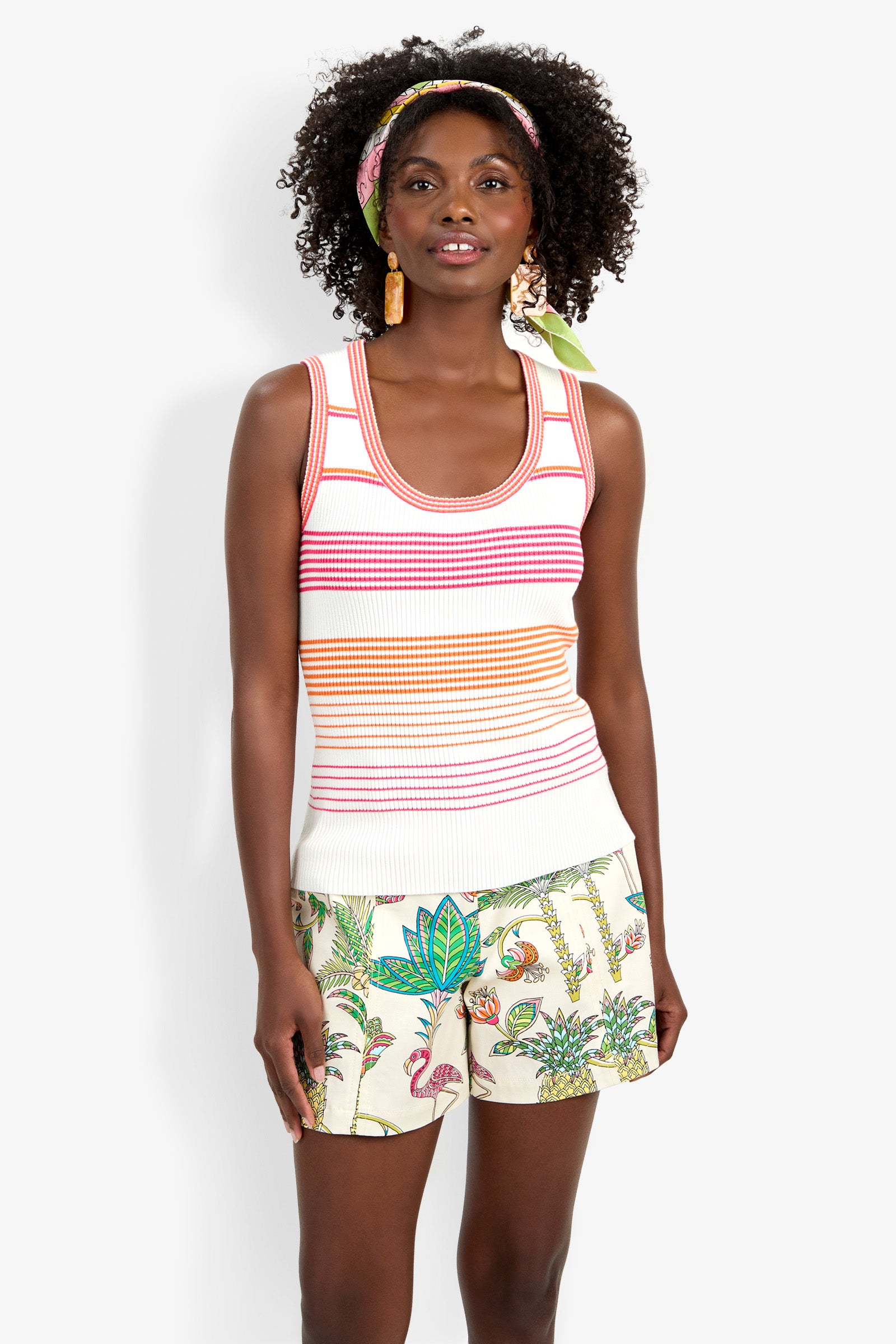 A woman with curly hair, a headband, and statement earrings smiles slightly while wearing the Paolini Tank and tropical-patterned shorts, standing facing forward.
