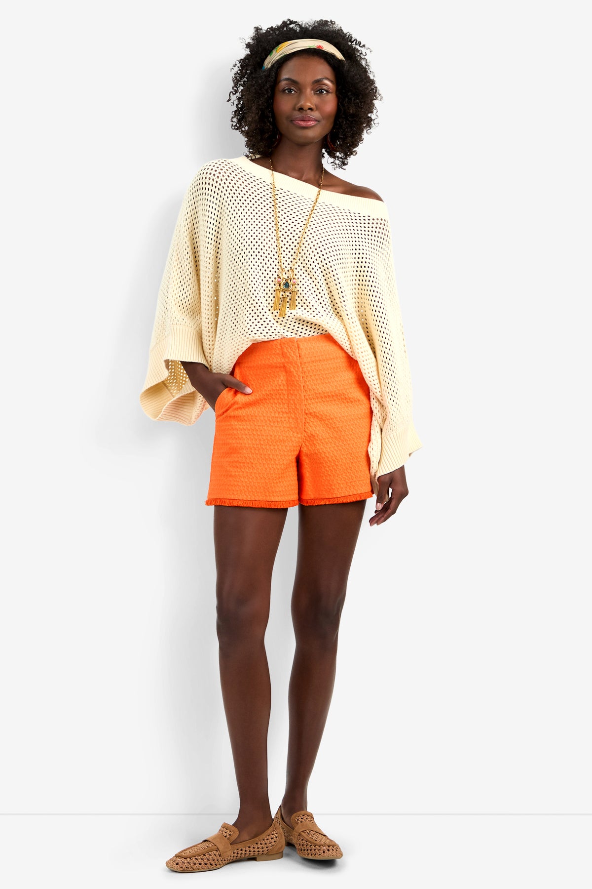 A woman wears the Horizon Pullover, bright orange shorts, tan woven sandals, a long necklace, and a yellow headband, standing against a plain white background with one hand in her pocket.