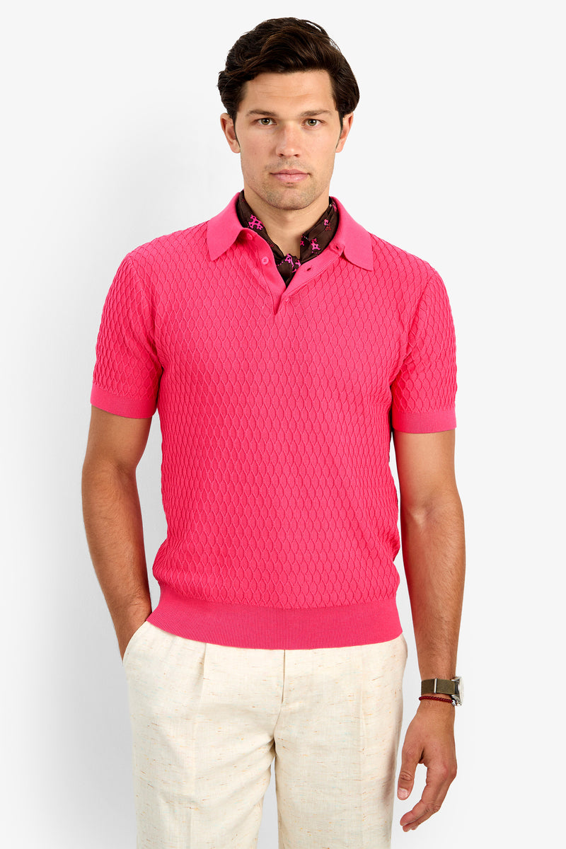 A man faces forward, wearing the HONALO SS POLO in Pitaya Pink, styled over a darker patterned shirt with off-white pants and a watch, set against a plain light background.