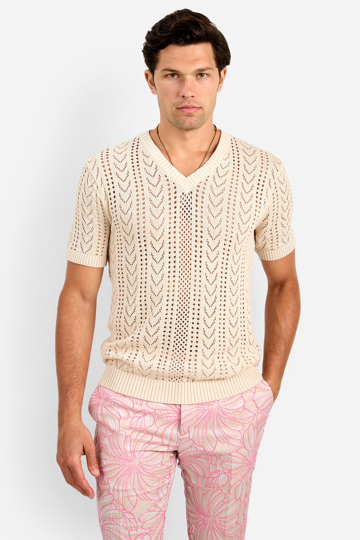 A man wears the KEANU CROCHET V-NECK, a cream short-sleeve cotton knit sweater with an openwork chevron pattern, paired with light pink floral pants, standing against a plain white background.