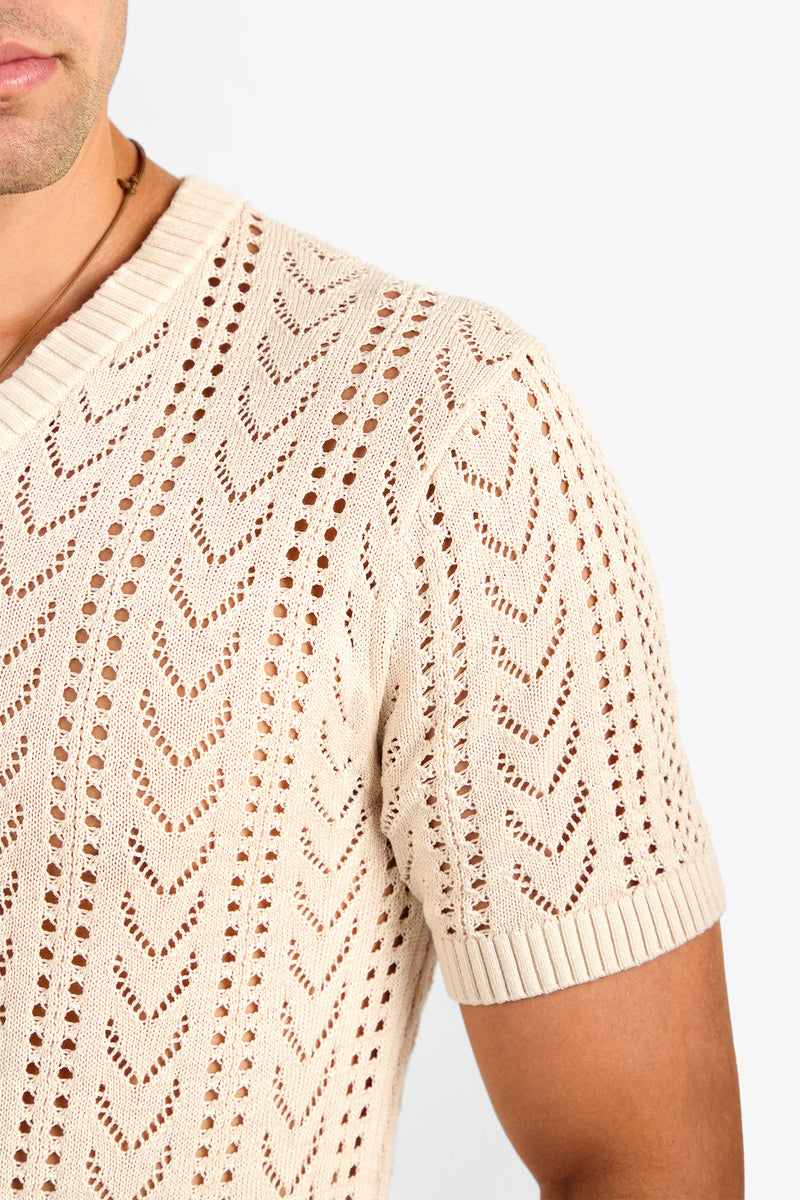 A close-up of a person wearing the KEANU CROCHET V-NECK, a beige short-sleeve knit shirt with chevron and eyelet patterns, ribbed collar, and sleeve cuffs. Only the torso, shoulder, and part of their face are visible.