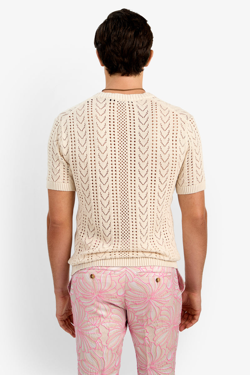 A man is seen from the back wearing the KEANU CROCHET V-NECK, a short-sleeve cream knit shirt with a chevron pattern, paired with light pink pants featuring a swirling abstract design.