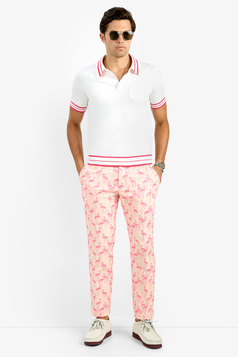 A man wearing sunglasses, a white polo shirt with pink trim, and Clyde Slim Trousers in light pink with a flamingo pattern stands with his hands in his pockets against a plain white background.