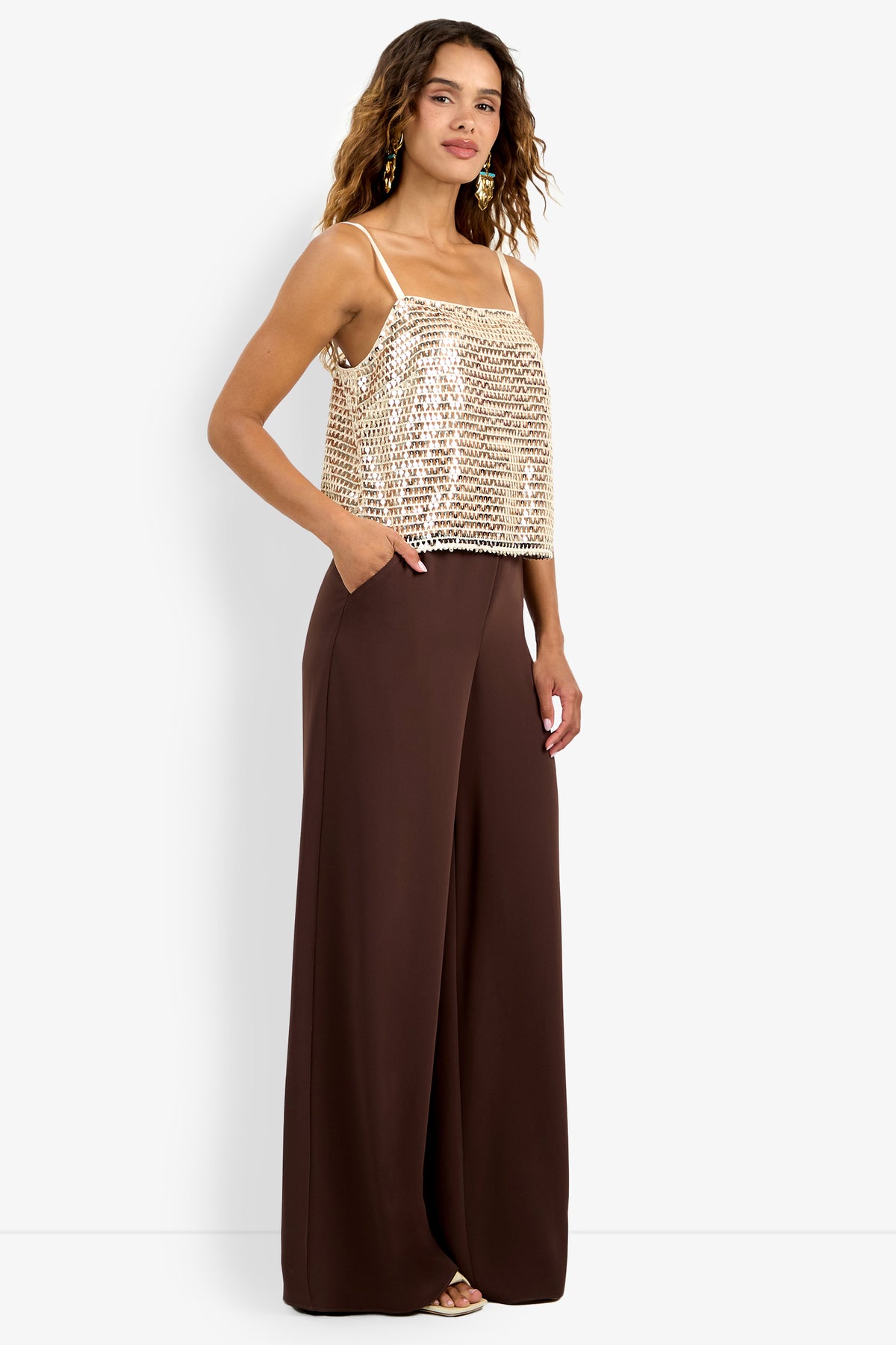 A woman in the Alannie Top, a sleeveless gold sequin crop top, and high-waisted wide-leg brown pants stands with one hand in her pocket, smiling and facing slightly to the side.