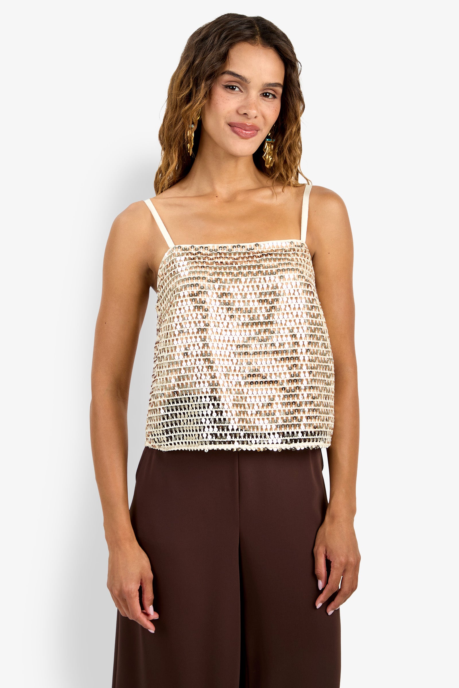 A woman with wavy brown hair wears the gold sequined Alannie Top with thin straps and brown wide-leg pants, standing against a plain white background.