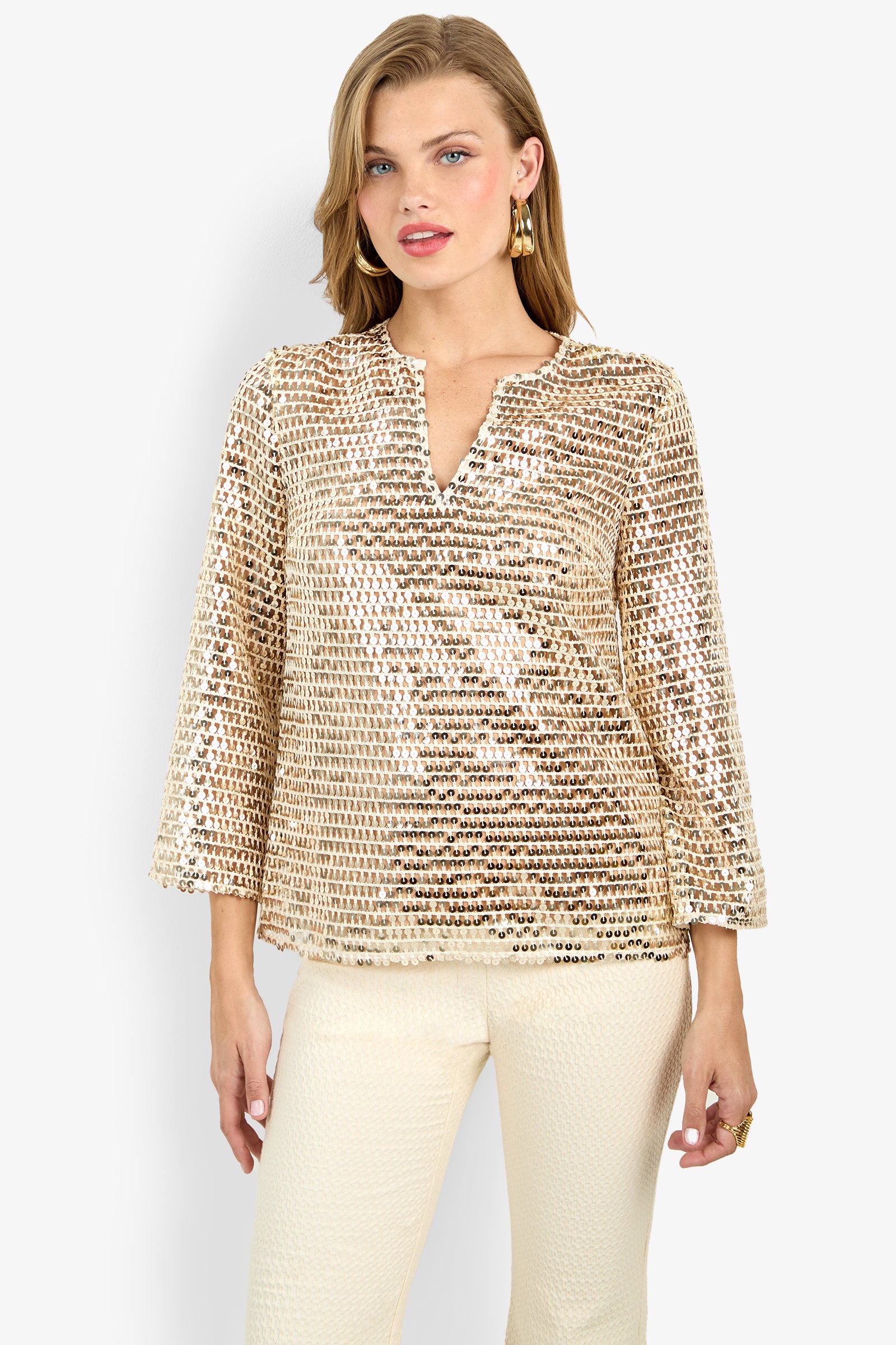 A woman wears the Mailani Top, a light gold sequin long-sleeve V-neck blouse, paired with cream textured pants against a plain light background.