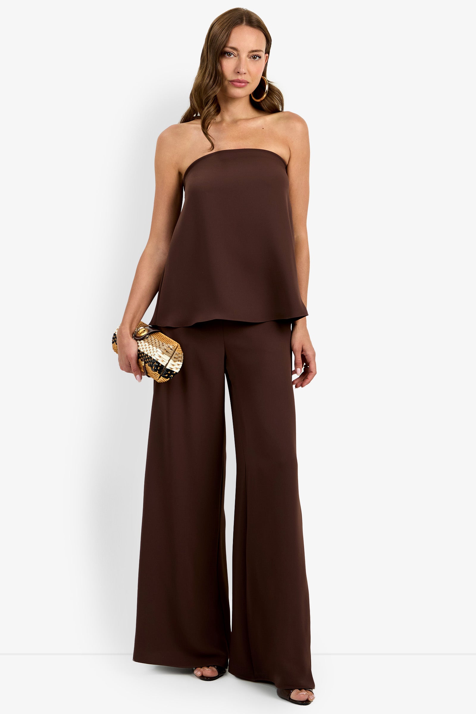 A woman wears the Hulla Top, a strapless, loose brown top paired with matching wide-leg pants. She accessorizes with hoop earrings, a small clutch, open-toed shoes, and her long wavy hair completes the look.