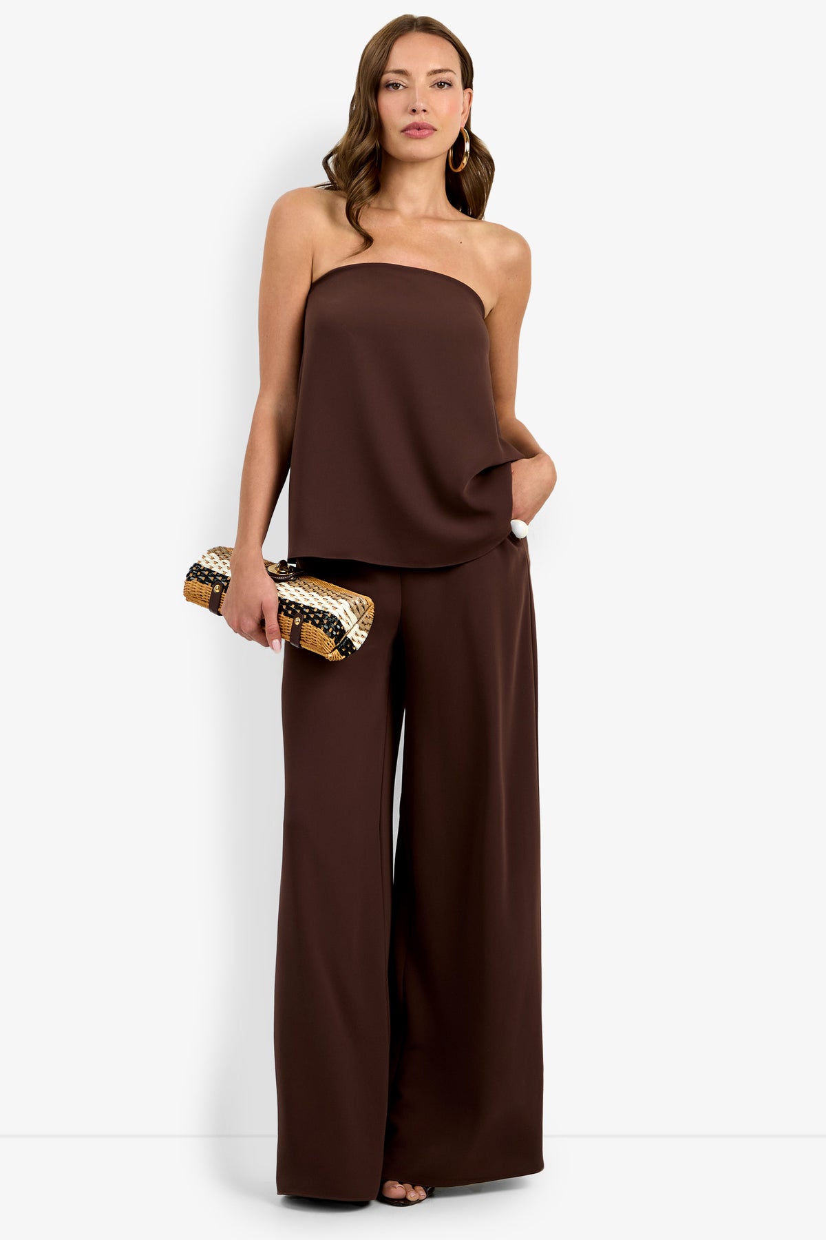 A woman stands against a white background wearing the Hulla Top, paired with wide-leg pants. She accessorizes with a gold and brown patterned clutch and brown hoop earrings, her hair styled in loose waves.