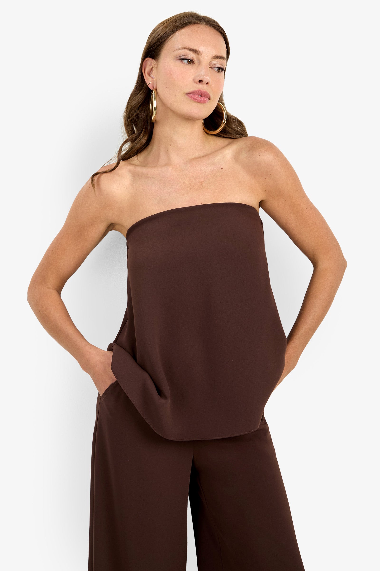 A woman with long brown hair and large hoop earrings stands against a white background, hands in her pockets, wearing the strapless dark brown Hulla Top and matching wide-leg pants.
