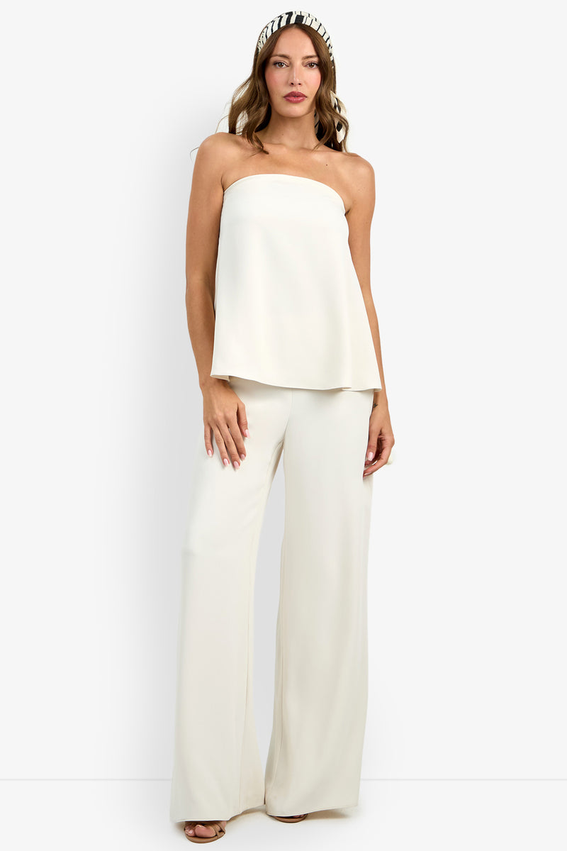 A woman poses against a plain white background wearing the Hulla Top, a strapless, loose-fitting white top paired with wide-leg white pants. She accessorizes with wavy hair, a headband, and open-toe heels.