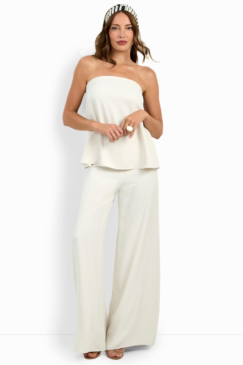 A woman poses against a plain white background in the Hulla Top, strapless and white, paired with wide-leg white pants and beige heels. She has loose, wavy hair styled with a black-and-white headband.