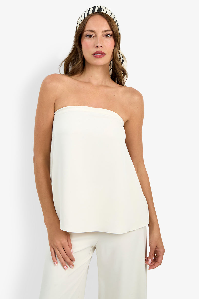 A woman with loose brown hair stands against a plain background, wearing the Hulla Top—an elegant strapless white top—paired with white pants and a black-and-white striped headband.