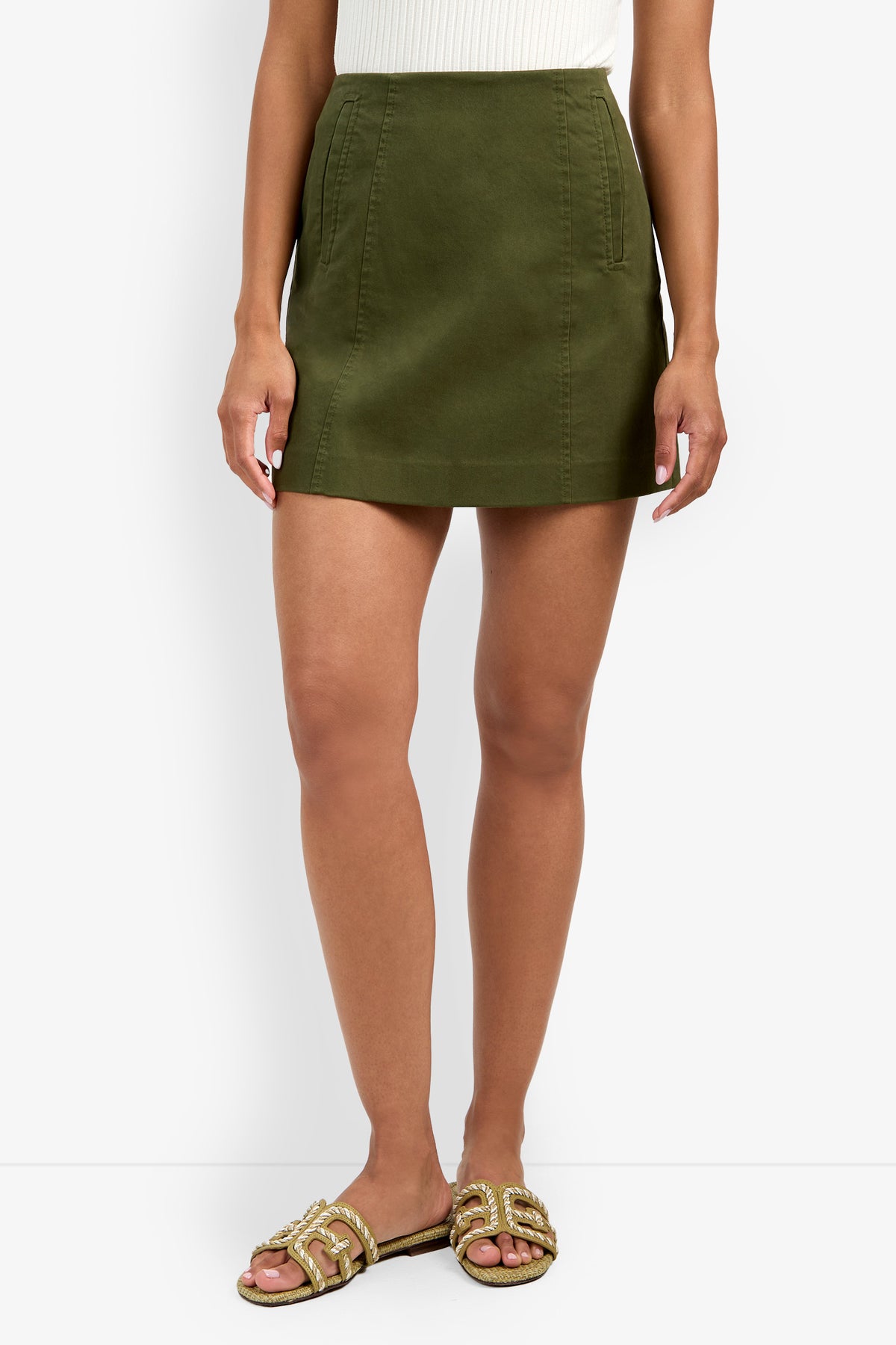 A person wears the Lavan Skirt—an olive green A-line mini—paired with a white sleeveless top and beige slide sandals with gold accents, standing against a white background.