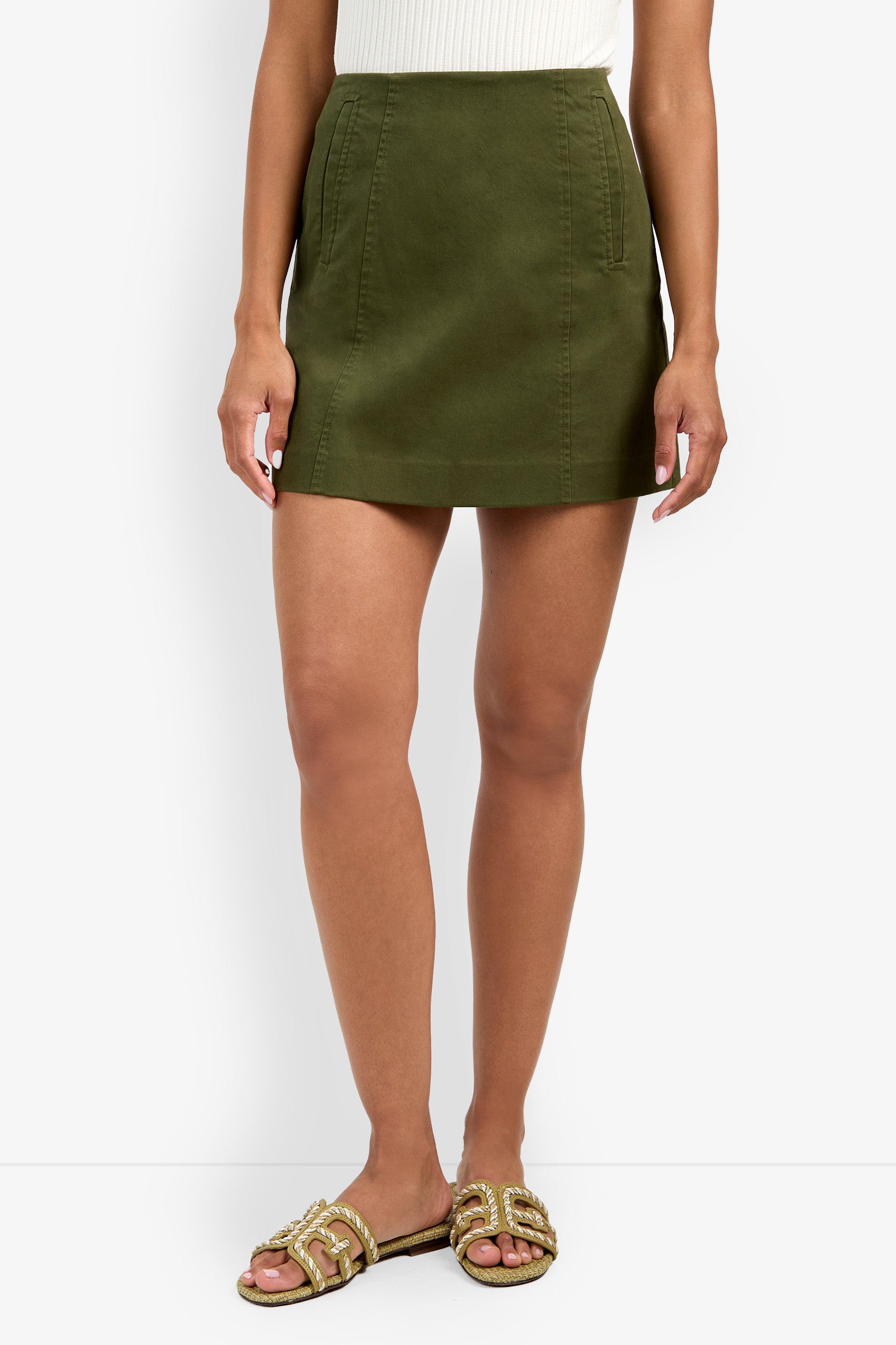 A person wears the Lavan Skirt—an olive green A-line mini—paired with a white sleeveless top and beige slide sandals with gold accents, standing against a white background.