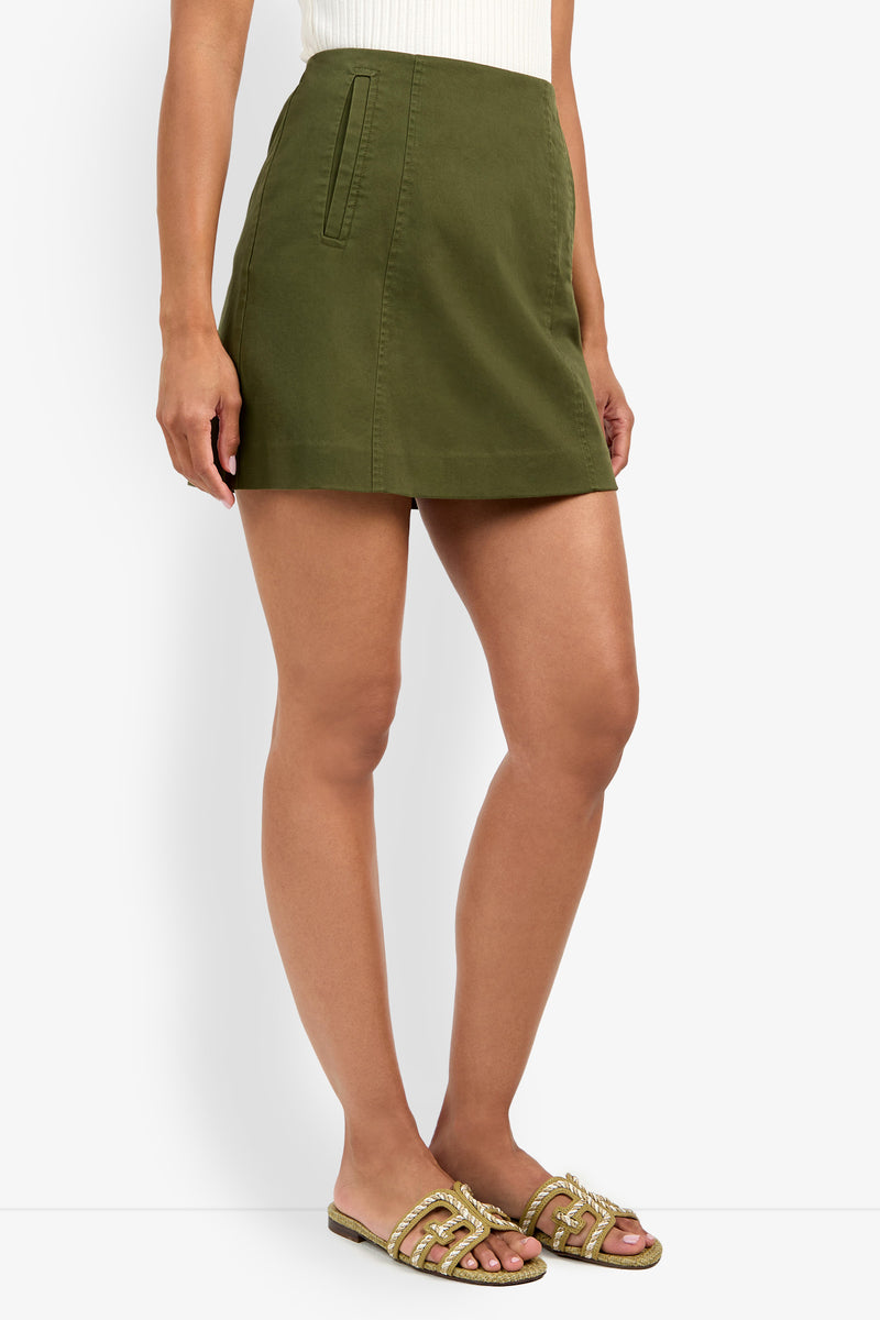 A person wearing a white ribbed top, the Lavan Skirt (an olive green mini skirt with side pockets), and gold embellished flat sandals stands against a white background.