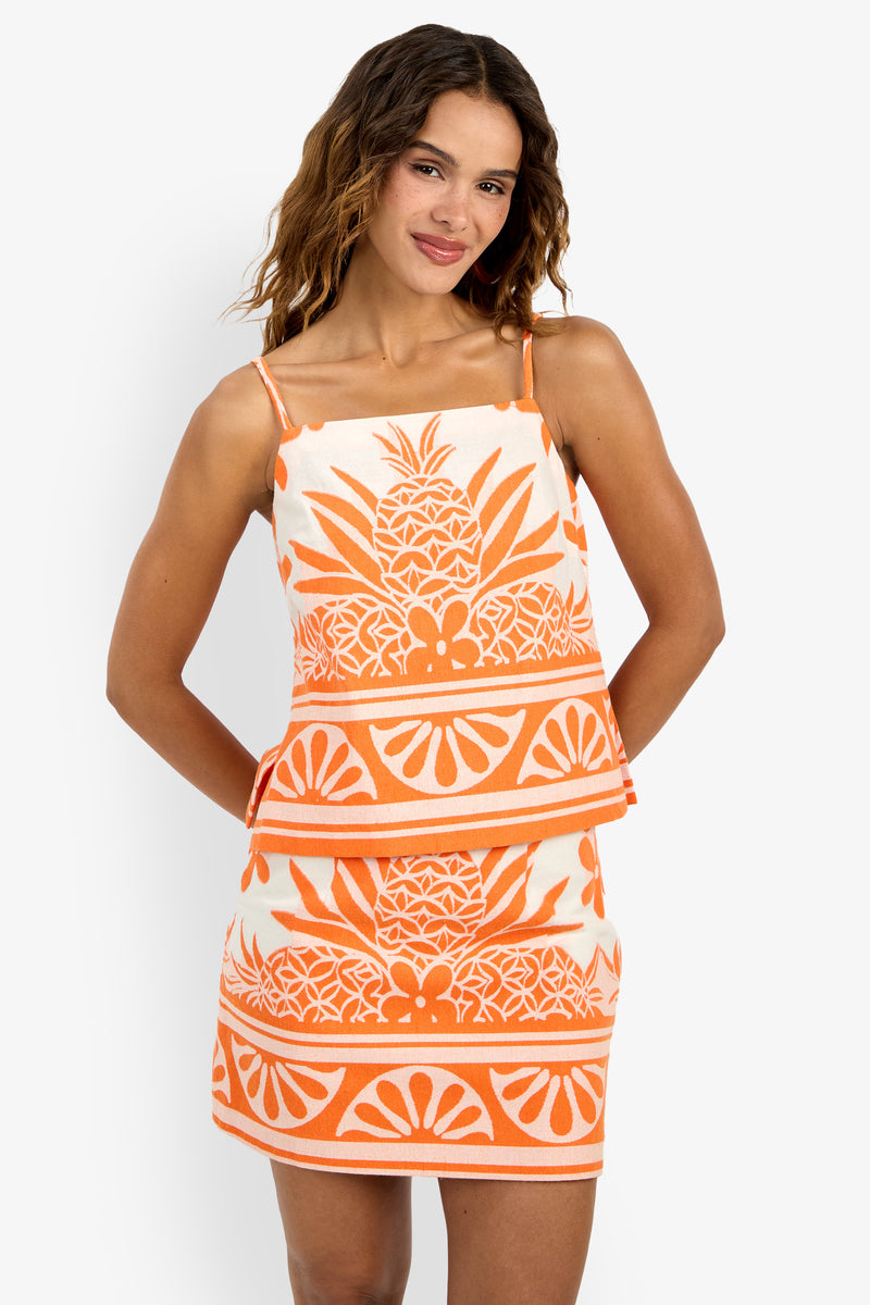A woman stands smiling with her hands behind her back against a white background, wearing the Marita Top—a sleeveless, orange and white piece featuring a tropical pineapple and geometric pattern.