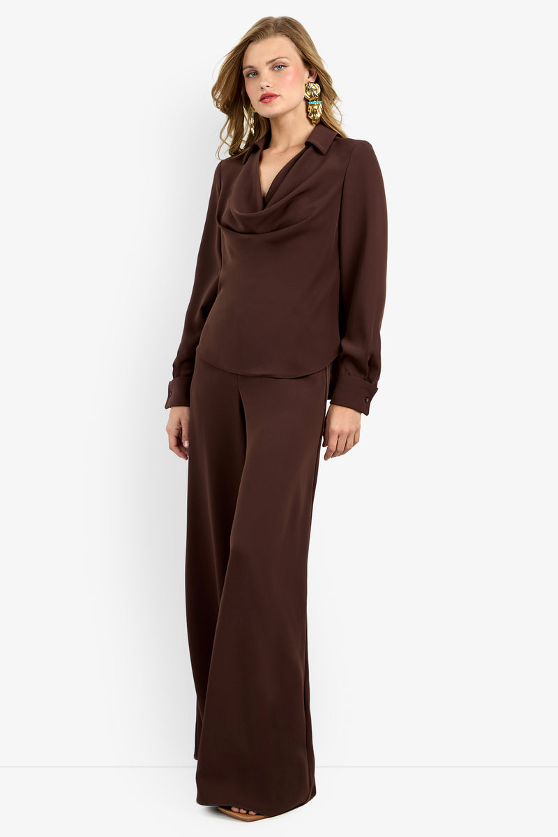 A woman stands against a white background wearing the Mistic Top, a dark brown long-sleeve cowl-neck blouse paired with matching wide-leg trousers and statement gold earrings.