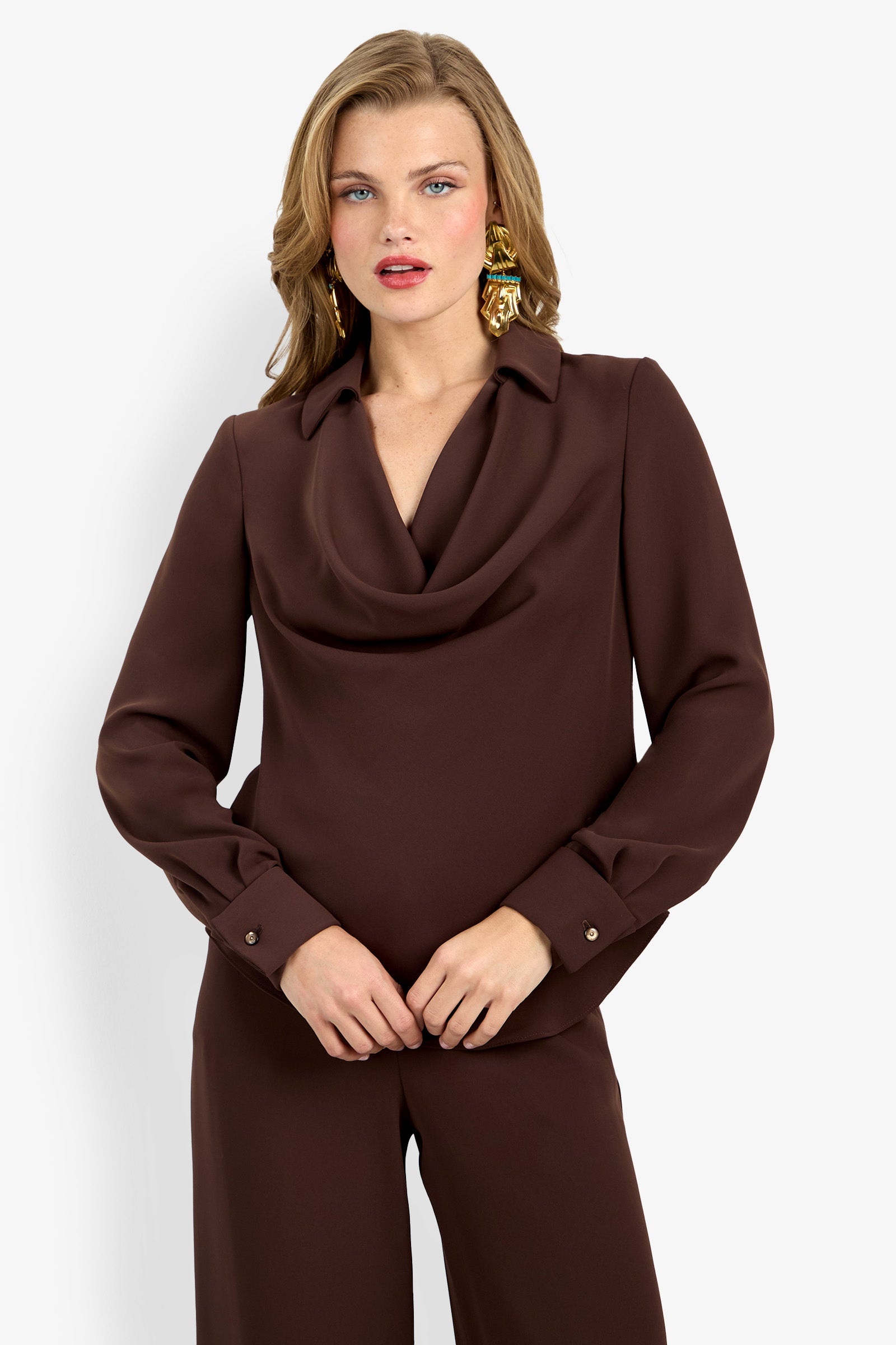 A woman wears the Mistic Top— a brown long-sleeve, draped-neck blouse— with matching brown pants and gold statement earrings, posing against a plain white background.