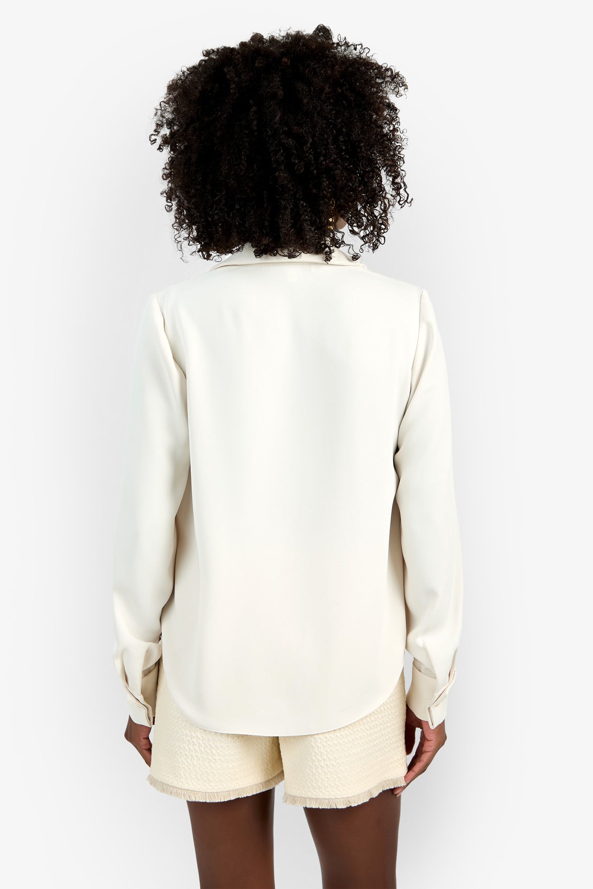 A person with curly hair, seen from the back, wears the Mistic Top—a long-sleeved white blouse—paired with textured cream shorts against a plain white background.