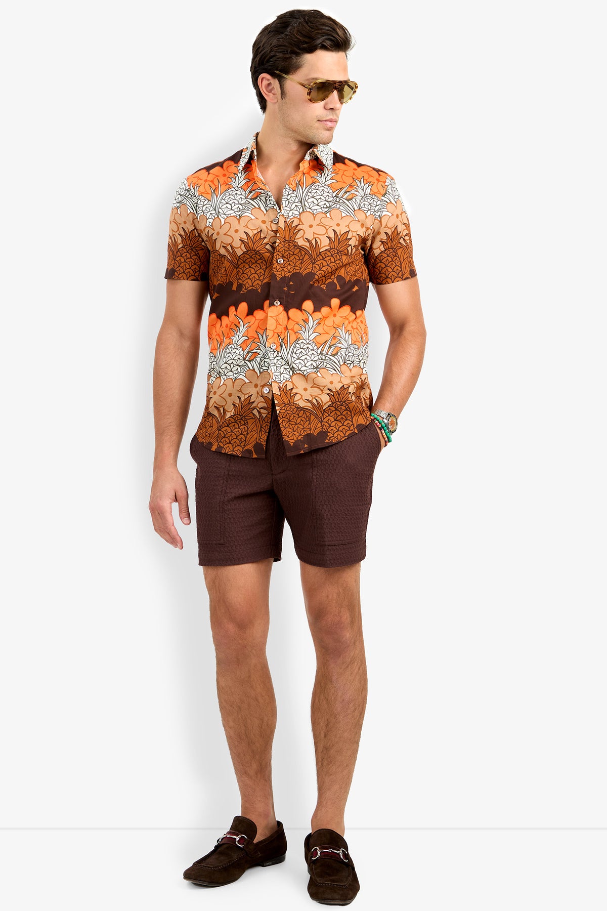 A man wearing sunglasses, the Slim Jim Shirt—a short-sleeve button-up with an orange, brown, and white floral pattern—dark brown shorts, and brown shoes stands with one hand in his pocket, looking to the side.