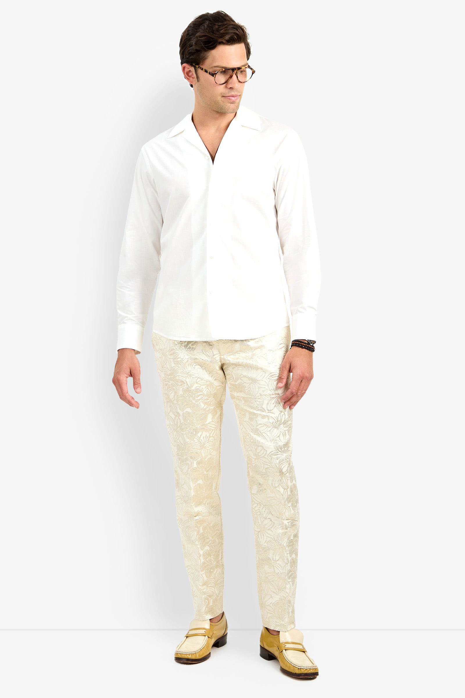 A man stands against a white background wearing the Able Shirt, patterned cream trousers, yellow loafers, glasses, and a dark bracelet, looking slightly to his right.
