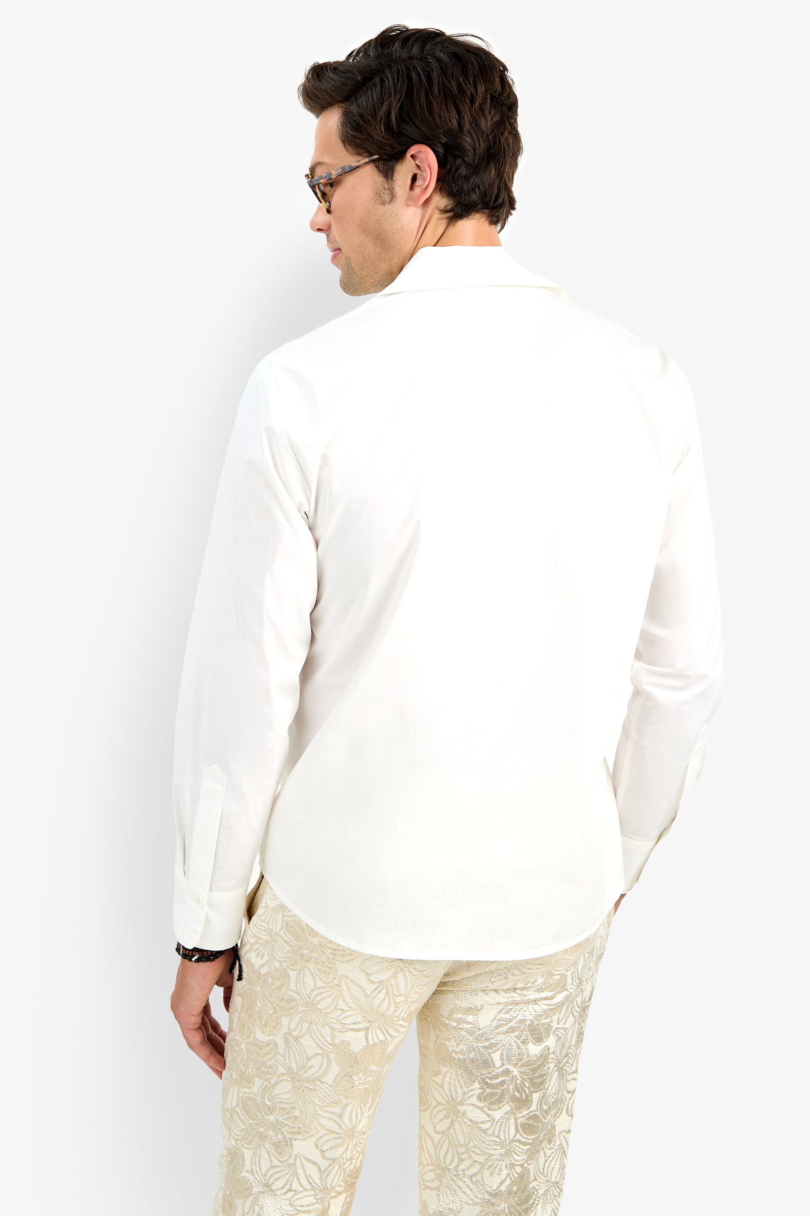A man with short dark hair and glasses is shown from the back, wearing the Able Shirt—a white long-sleeve button-down—paired with light floral pants, standing against a plain white background.